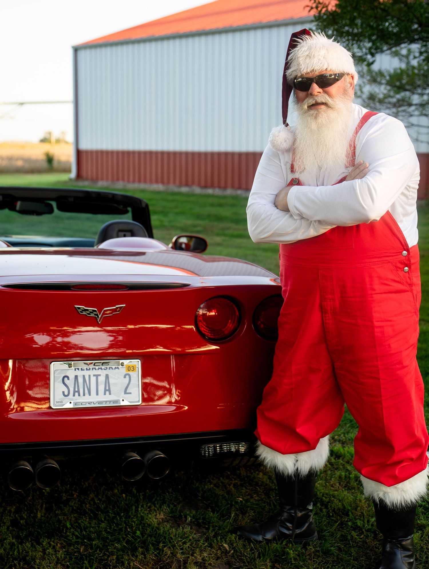 Santa Claus leaning against a red Corvette sports car with a SANTA 2 license plate outdoors.