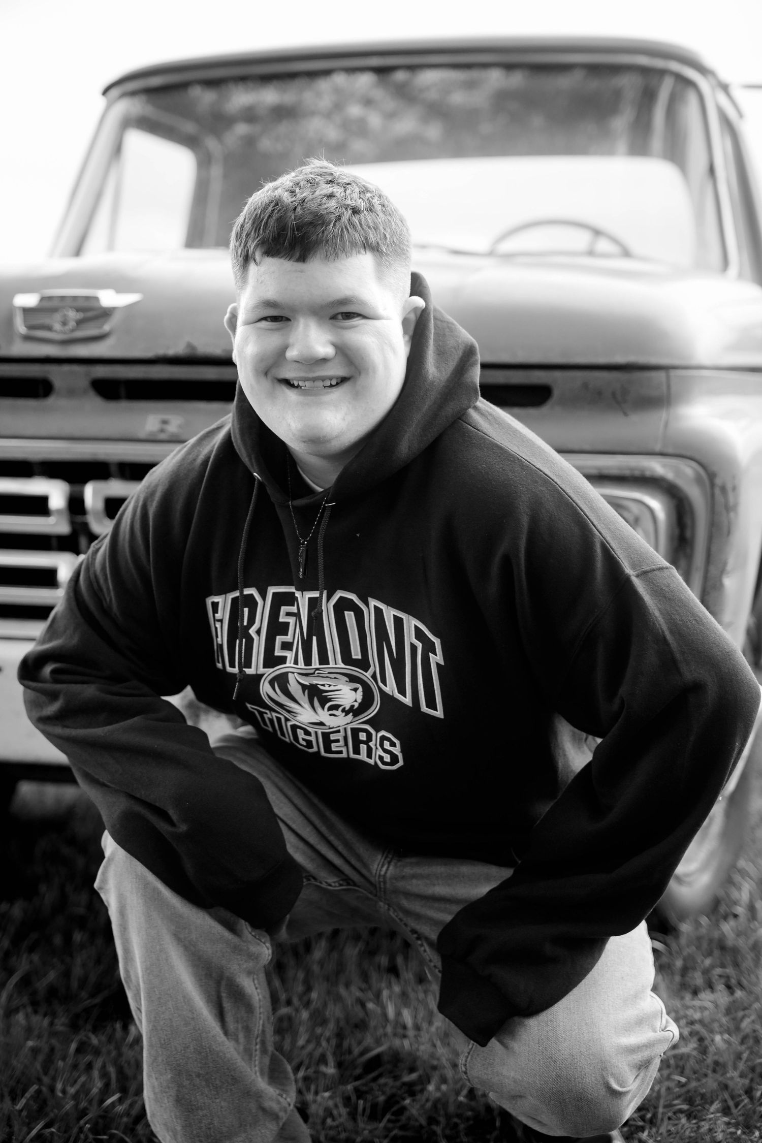 Smiling young man in a Fremont Tigers hooded sweatshirt kneeling in grass in front of a vintage pickup truck in a black and white photo.