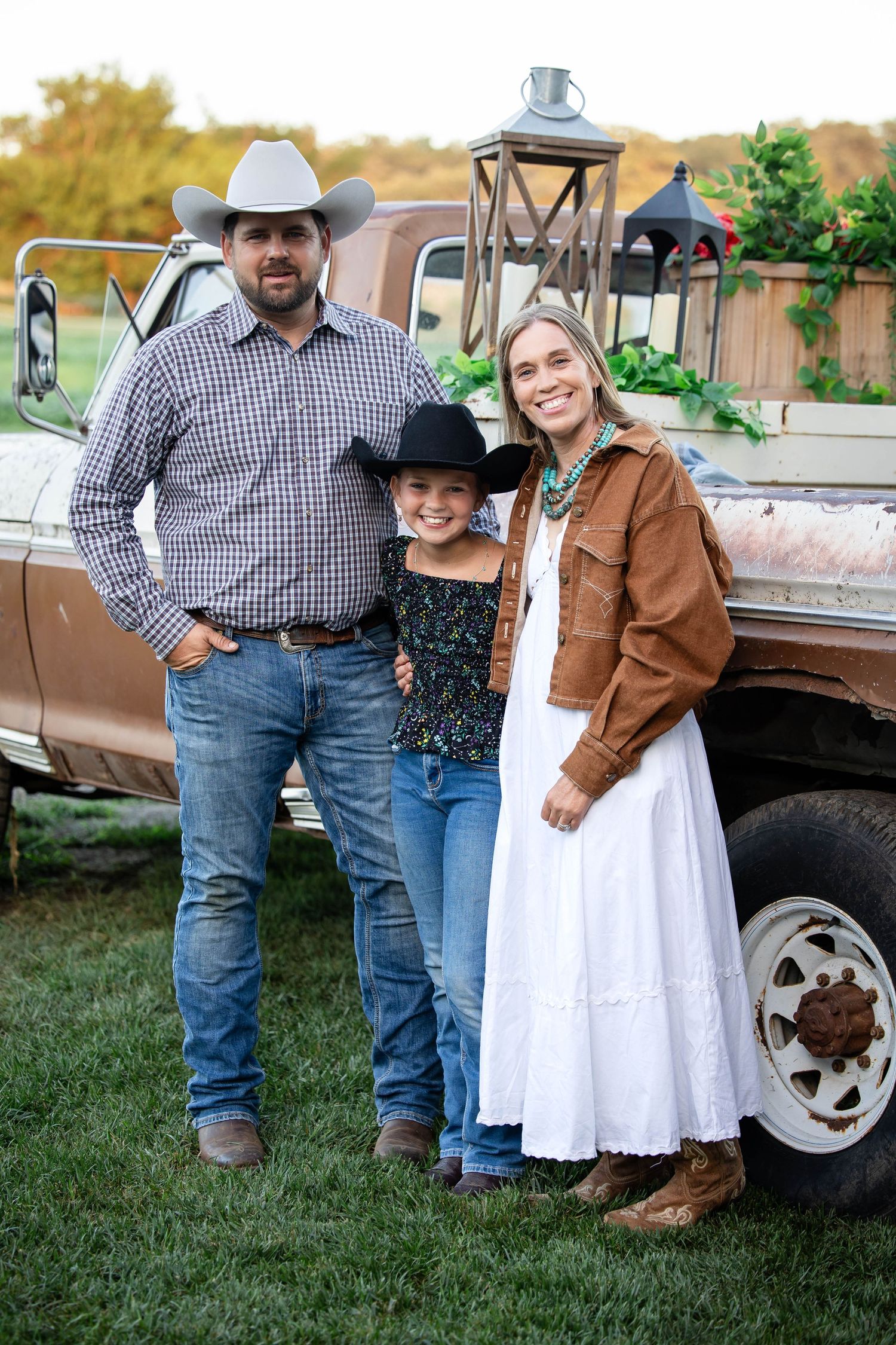 Family of three in western wear pose by a vintage truck at a fall farm event, wearing cowboy hats and boots.