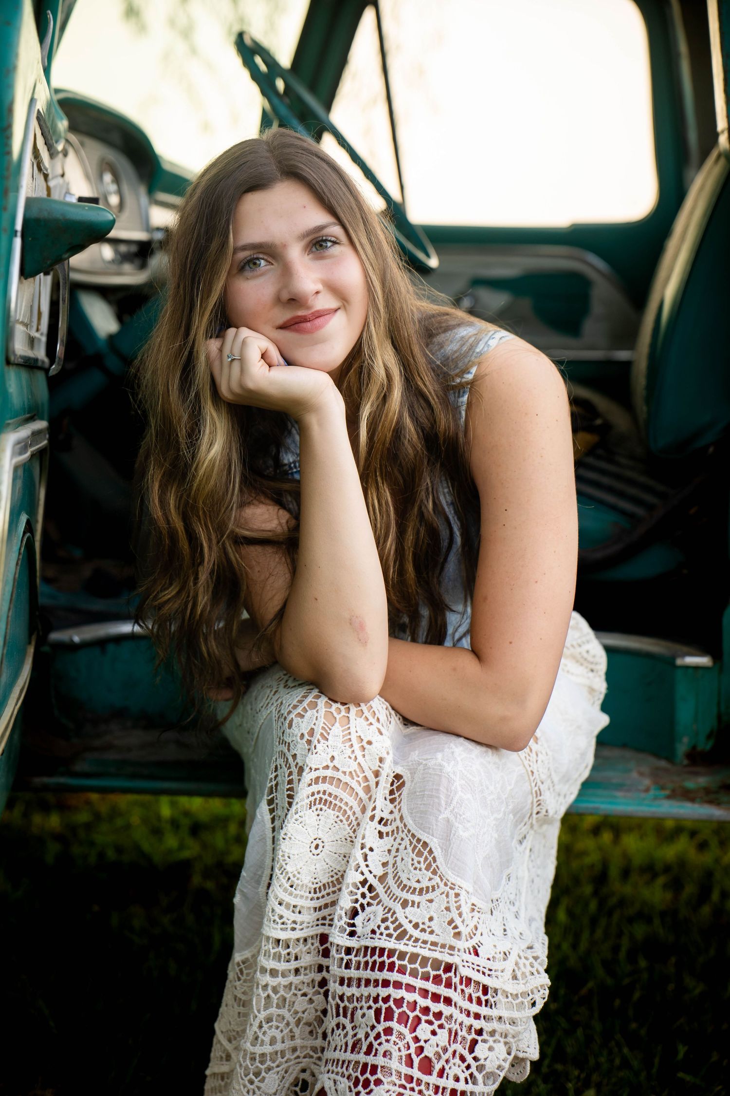 Smiling teenage girl with long brown hair sitting in vintage green truck doorway with lace skirt.