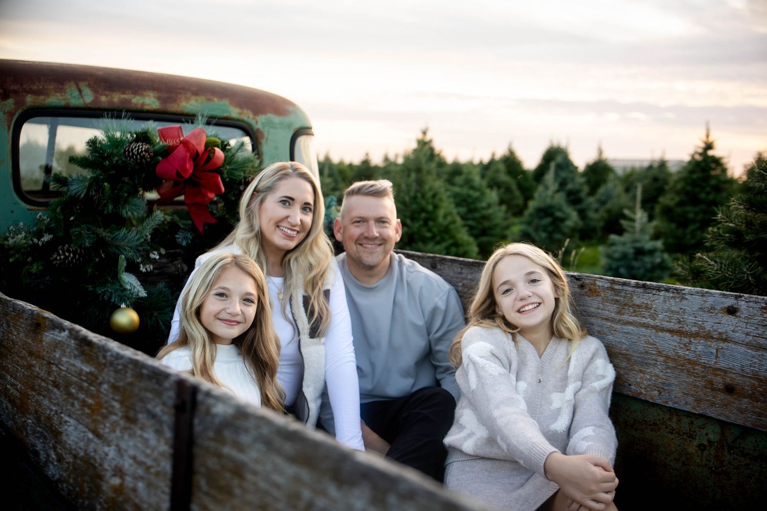 Smiling family of four sits in a vintage truck bed decorated with a red poinsettia wreath, with a Christmas tree farm behind them at sunset.
