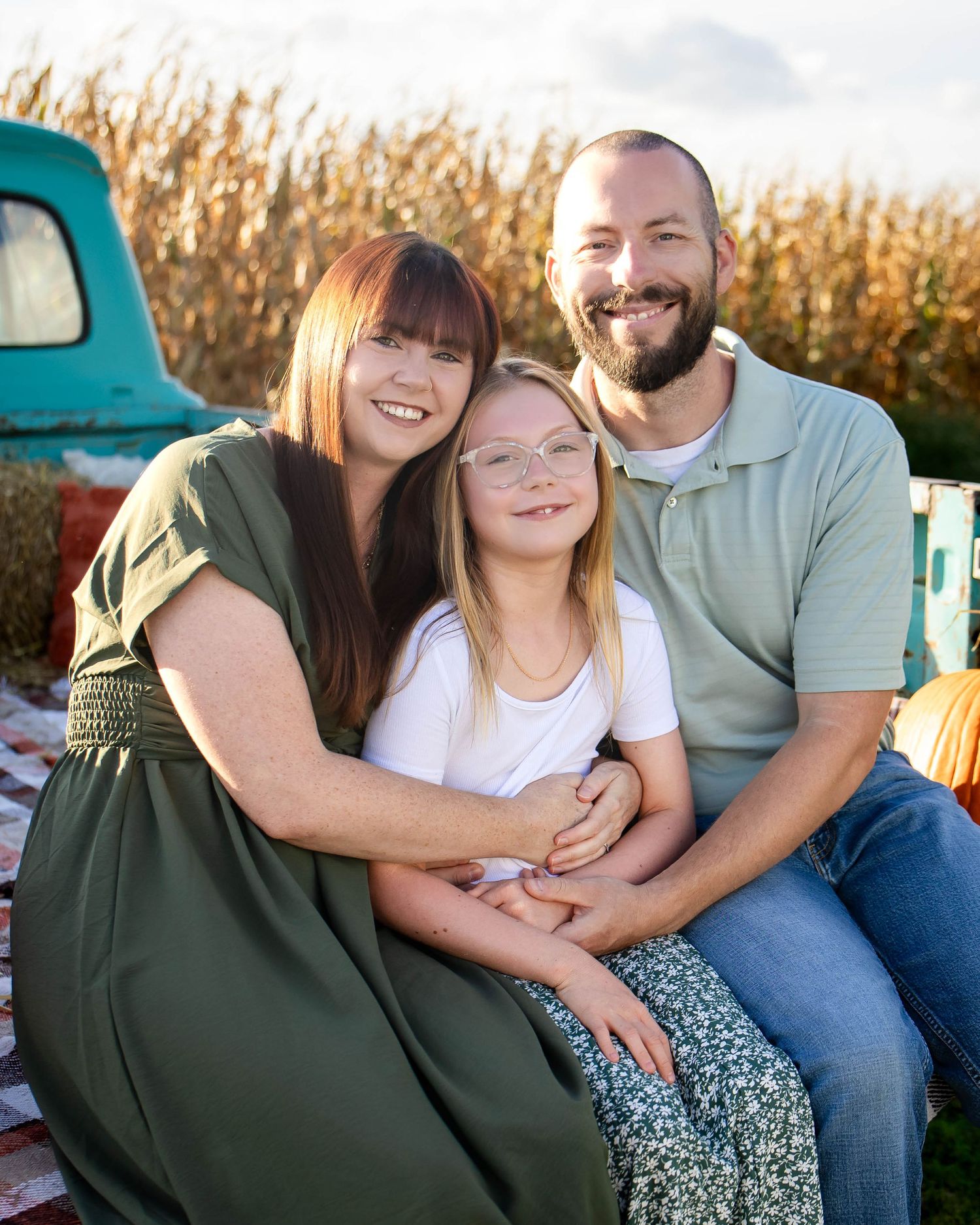 Happy family of three sitting together on vintage truck bed at fall corn field photo session.
