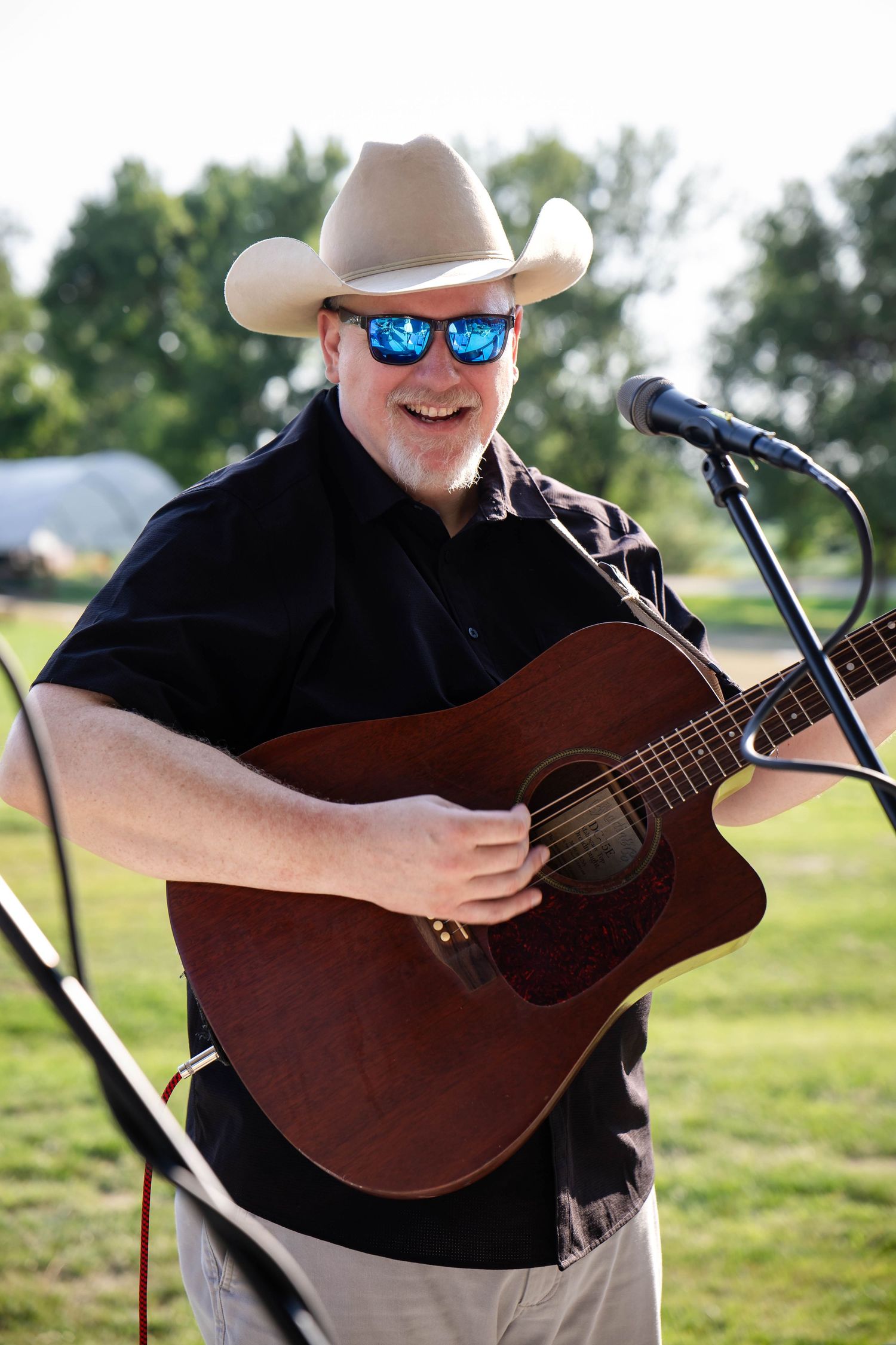 Male country musician in cowboy hat and sunglasses plays acoustic guitar at outdoor performance.