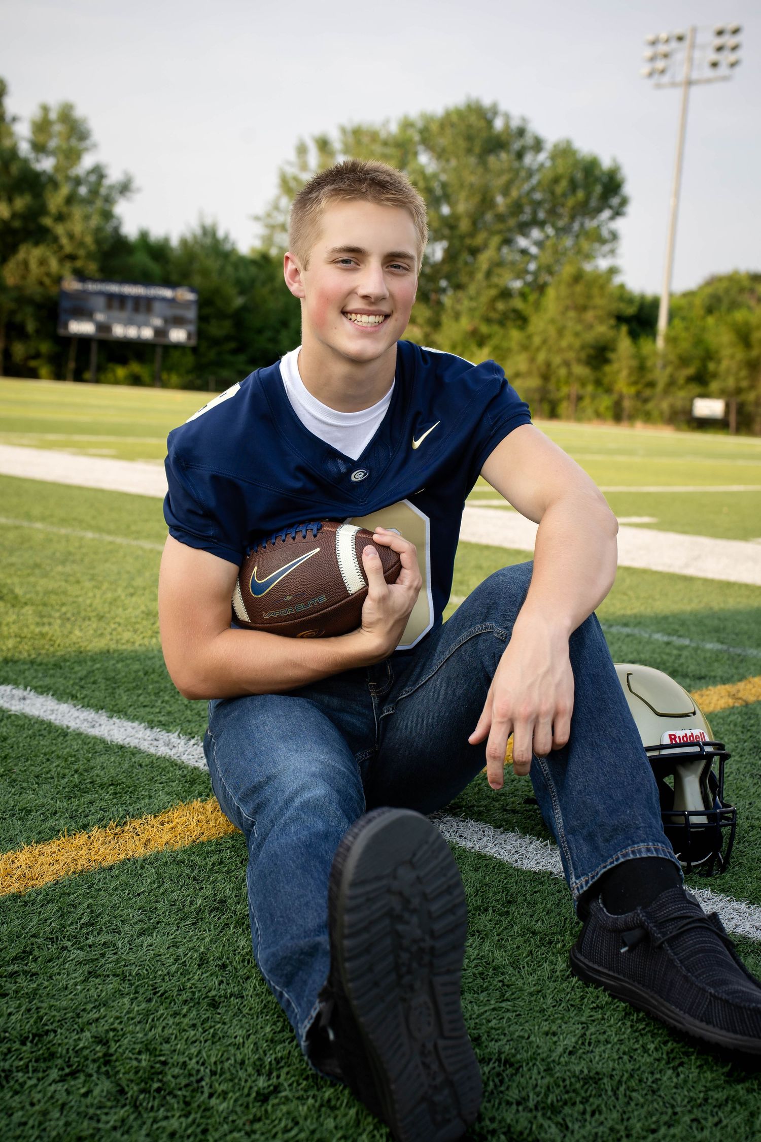 Smiling teen football player in navy jersey sitting on field holding football with helmet nearby.