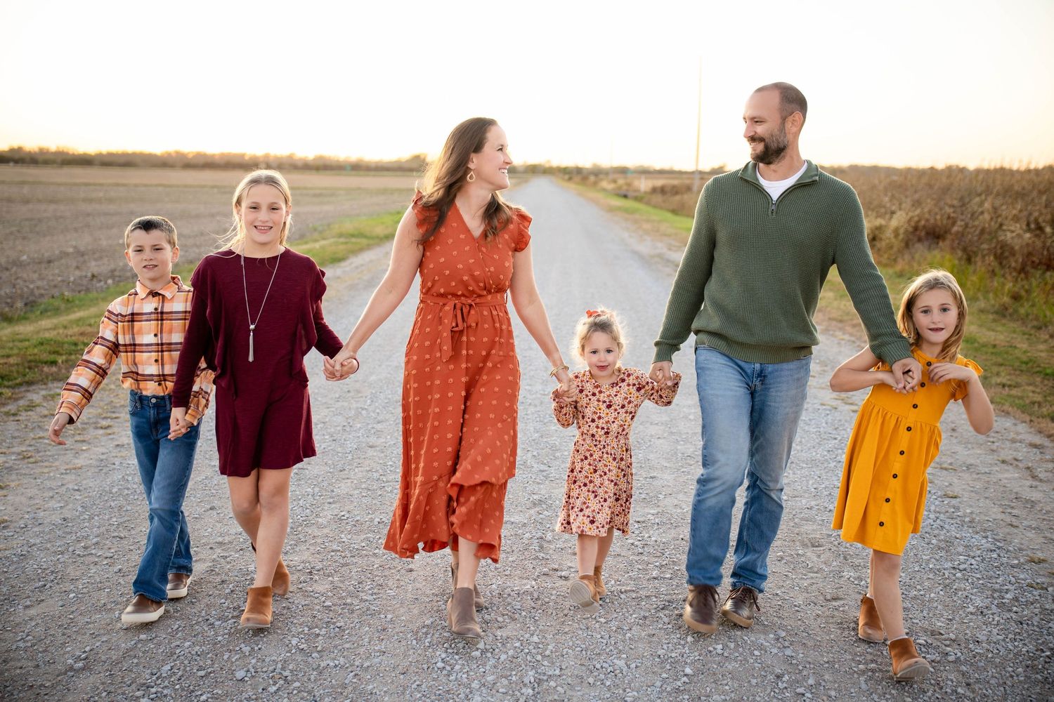 Family of six holding hands walking down rural dirt road during golden hour fall photo session.