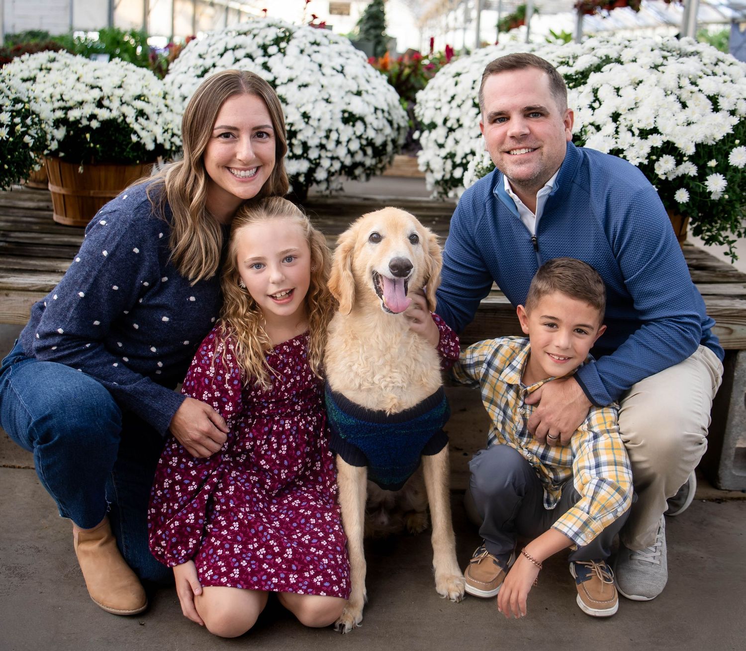 A smiling family of four poses with a happy golden retriever in front of blooming white chrysanthemums at a garden center.