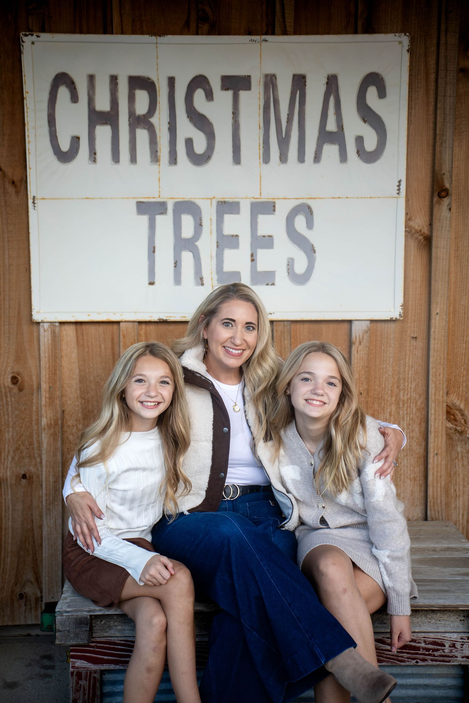 A smiling woman sits with two young girls in front of a rustic Christmas Trees sign on a weathered wooden wall.