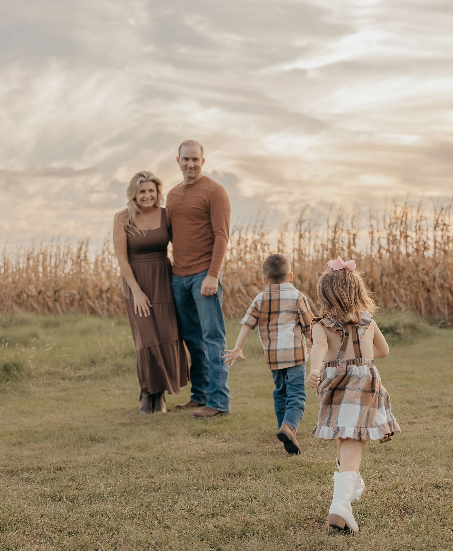Parents stand in a golden cornfield as two young children in plaid run toward them under a dramatic sky.
