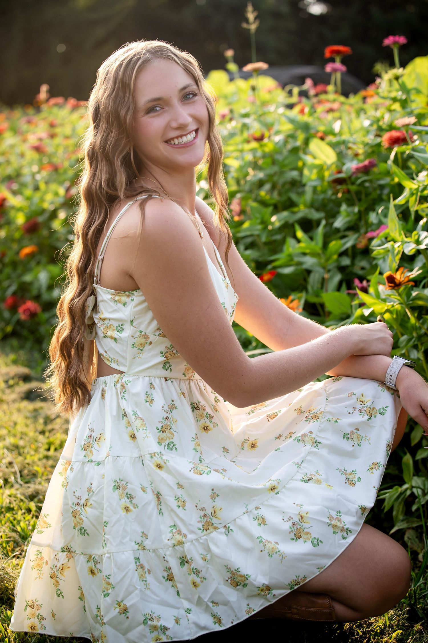 Smiling teenage girl in floral sundress sitting among colorful zinnia flowers in a sun-drenched garden.