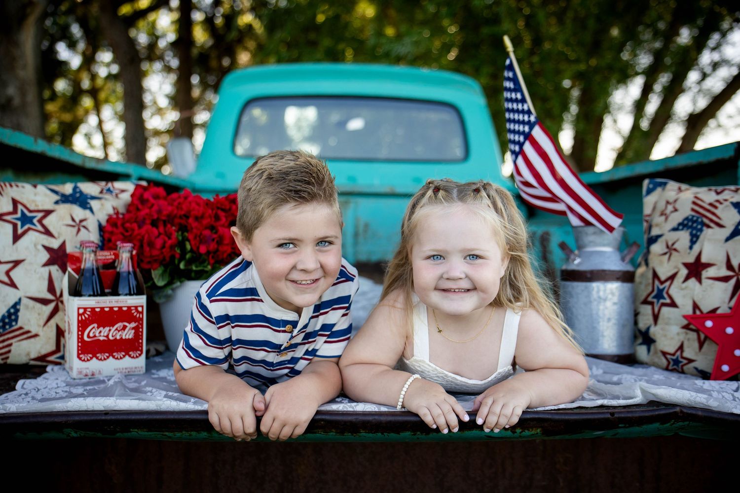 Two young children smiling in back of teal vintage truck with American flag and Coca-Cola decor.