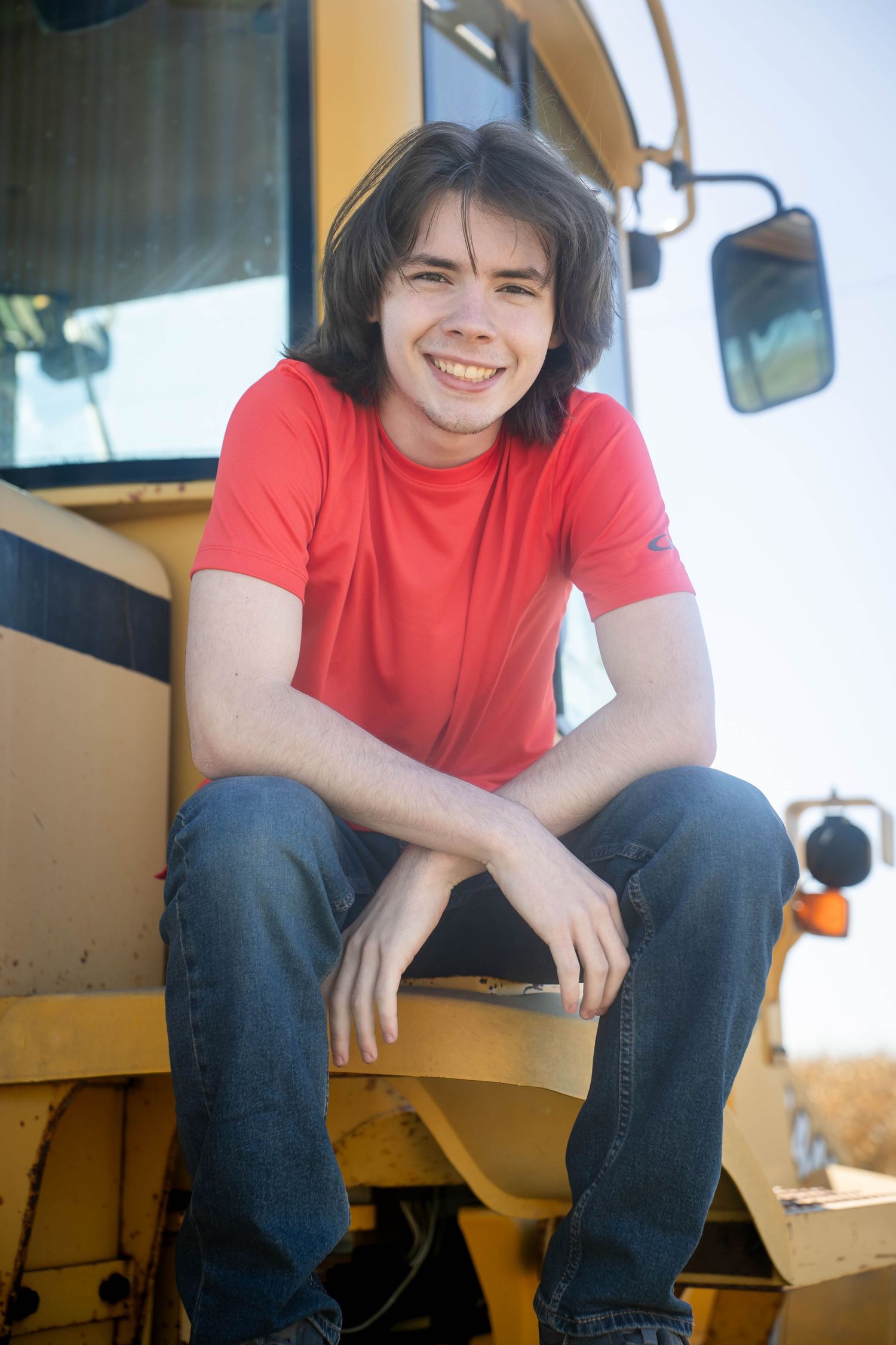 Smiling young man in a red t-shirt and jeans sits on the step of a yellow farm vehicle on a sunny day.