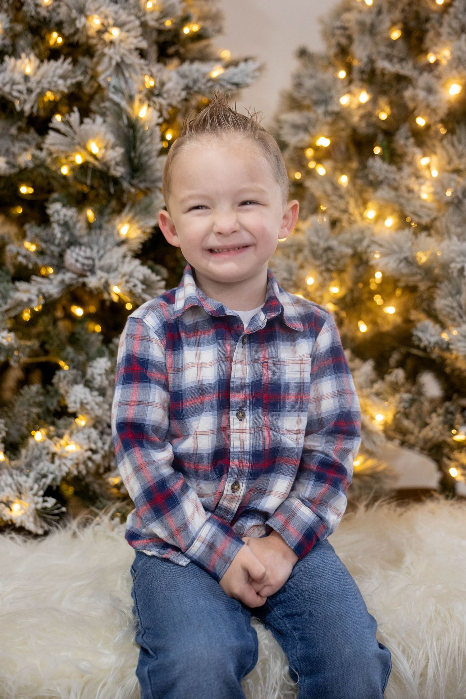 Smiling young boy in a plaid flannel shirt and jeans sits on a white fur rug in front of two lit, frosted Christmas trees.