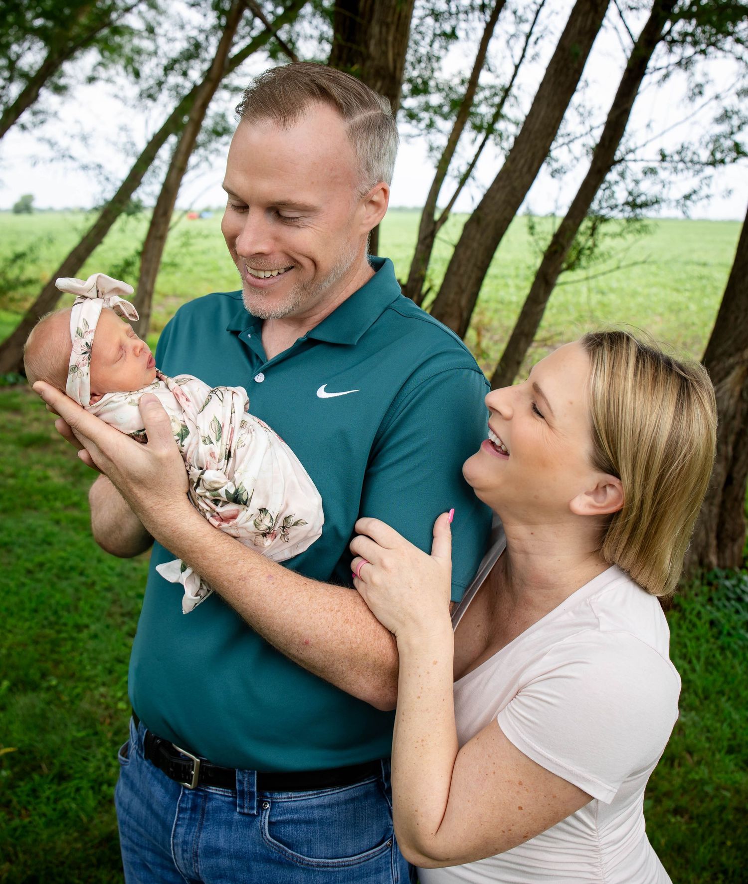 Proud parents smiling and cradling swaddled newborn baby outdoors in green summer setting.