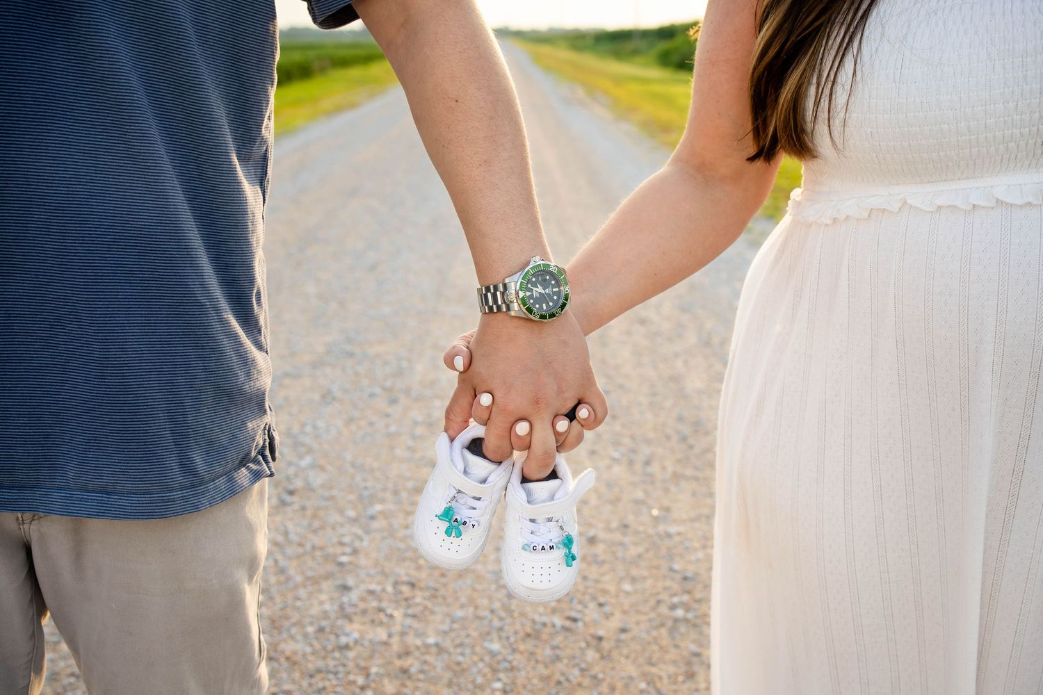 Couple holding hands and tiny baby shoes together on a gravel road for pregnancy announcement.