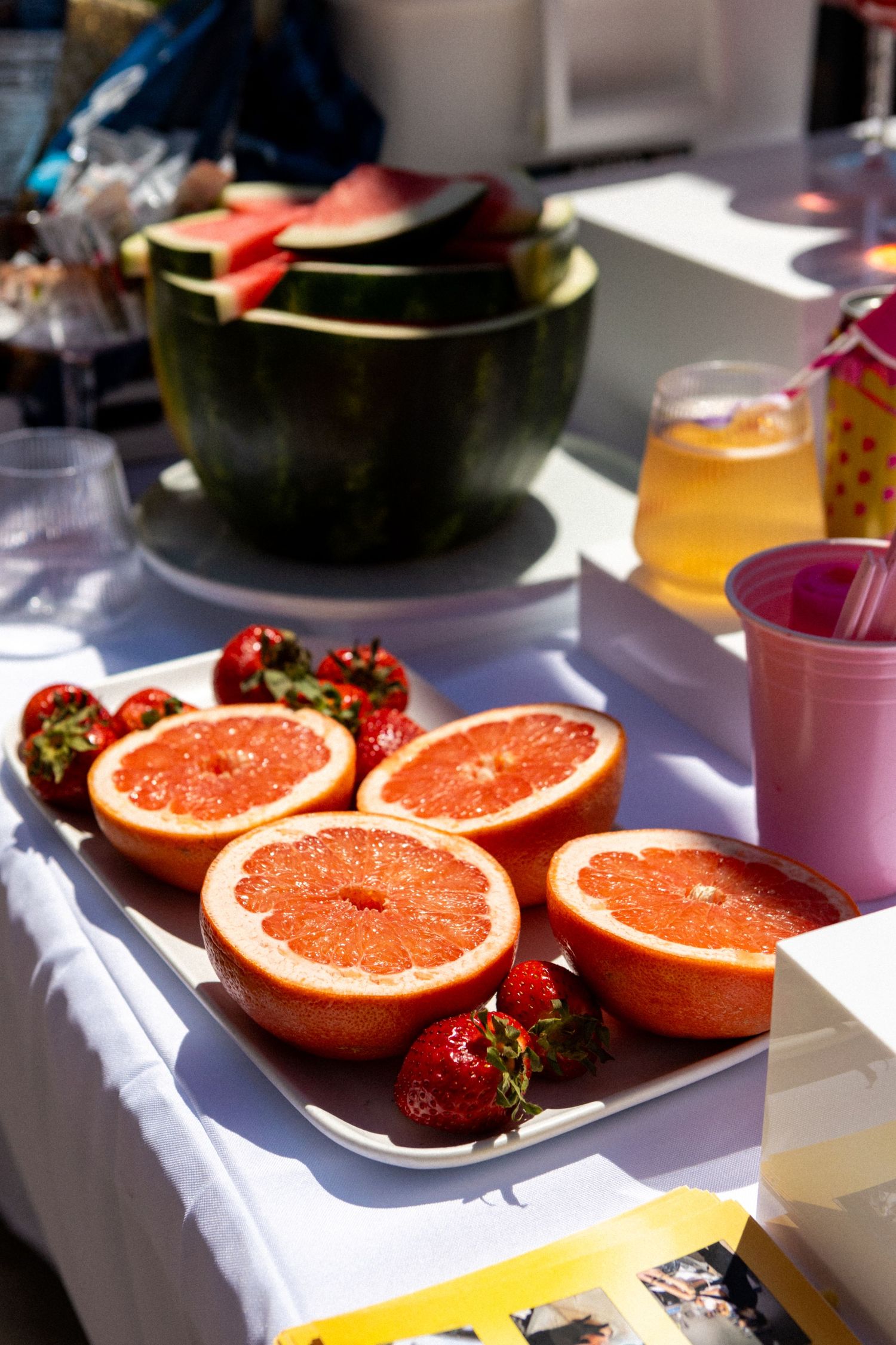 Fresh halved grapefruit and strawberries on white platter next to colorful drinks in sunlit outdoor setting.