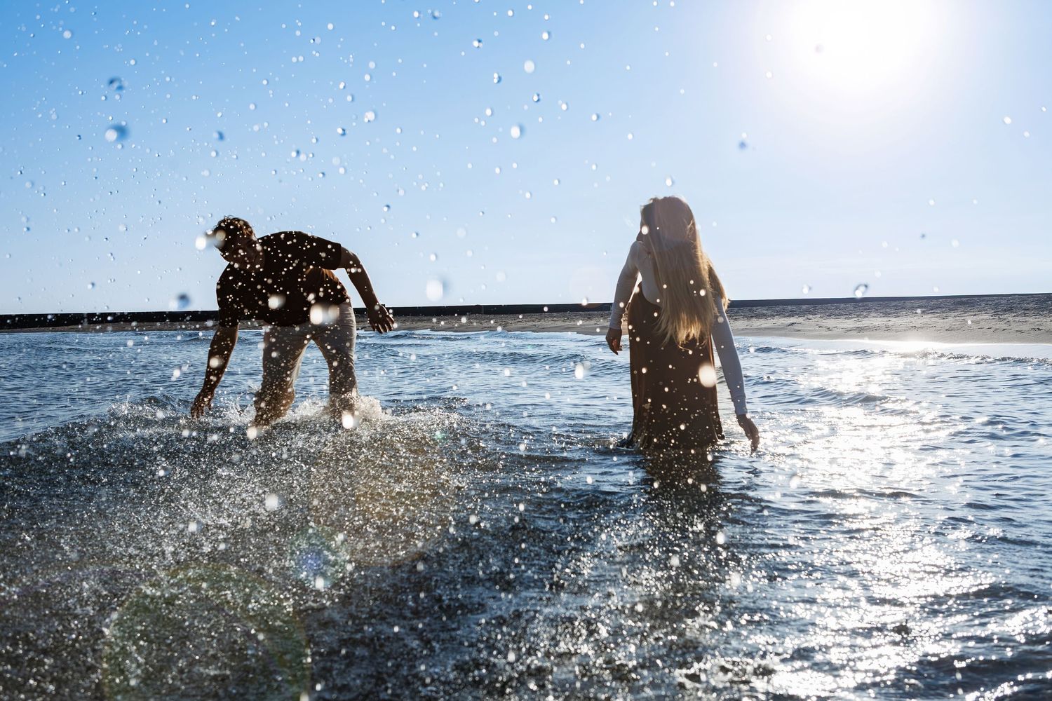 Silhouettes of people and dogs playing in ocean waves at sunset creating splashing water reflections in golden light.