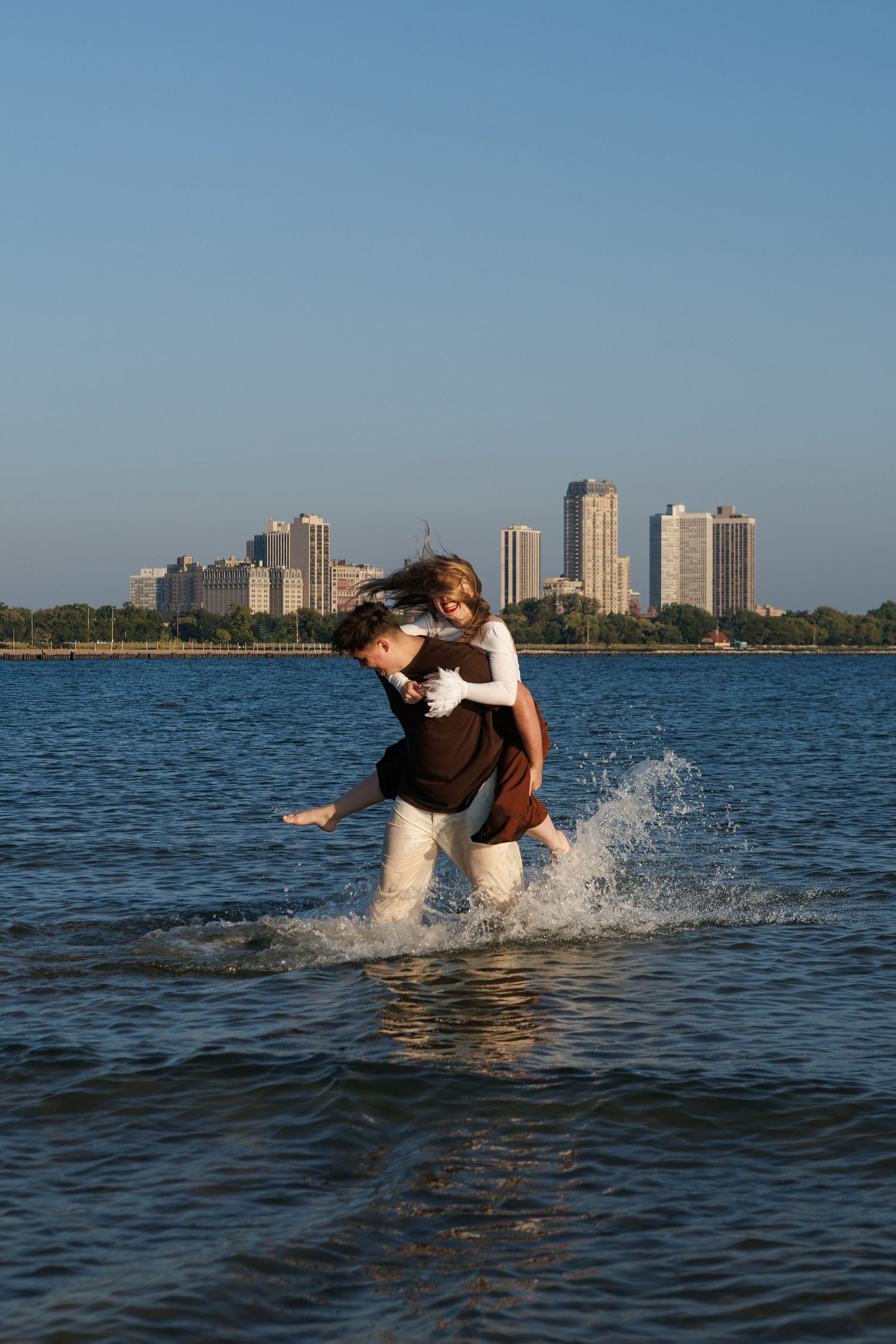 Two people splash and fall into Lake Michigan with Chicago's skyline visible in the background during sunset.