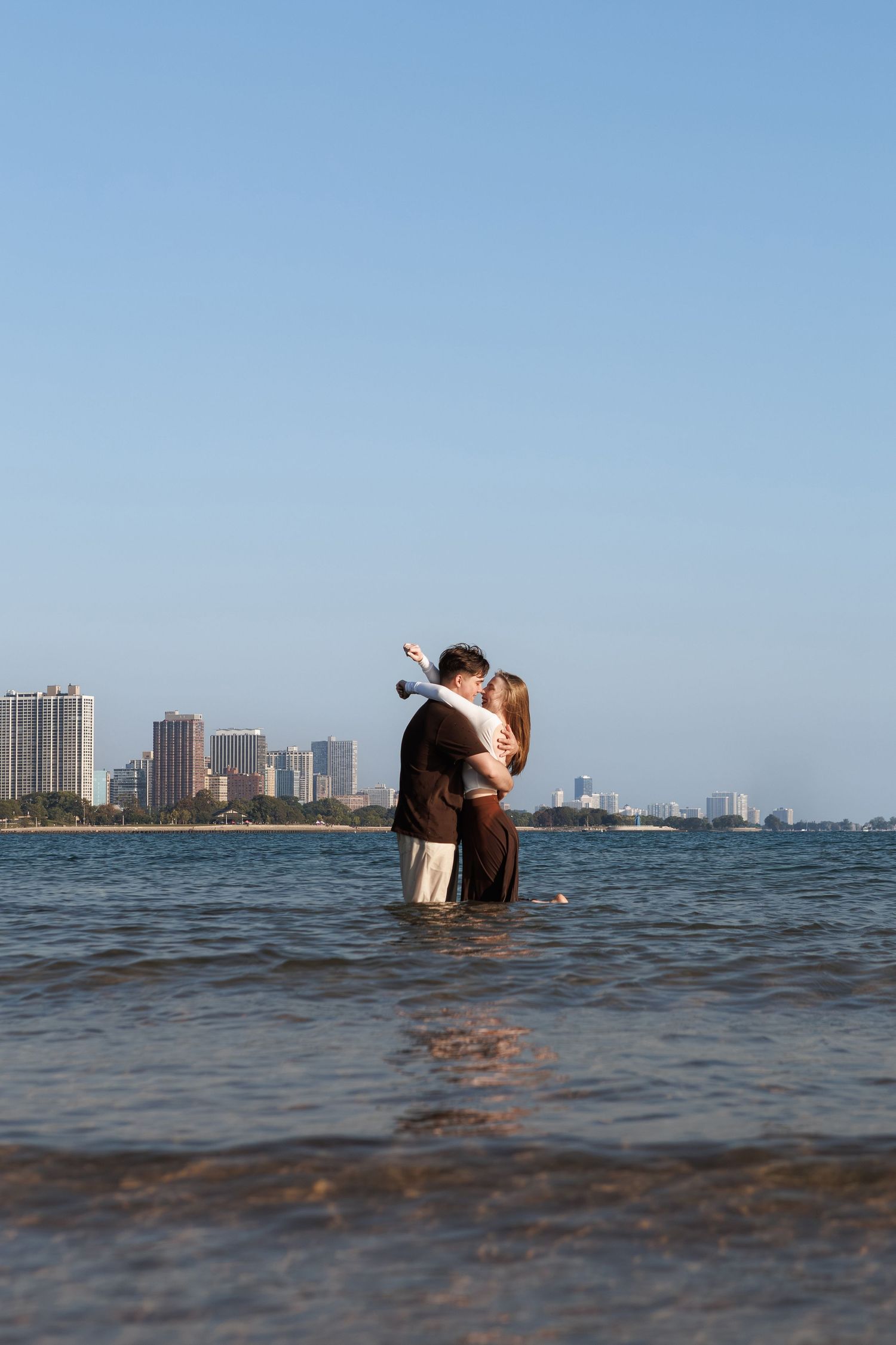 A person stands in shallow ocean water with a city skyline visible in the distance.