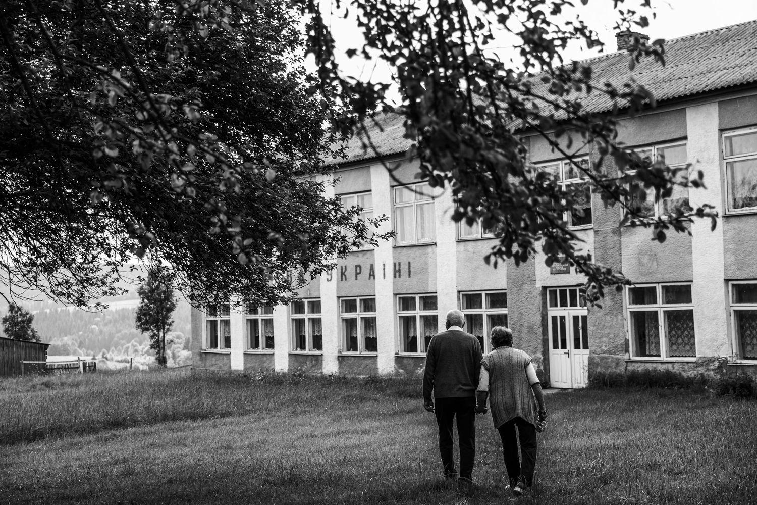 An elderly couple walks together on a grassy lawn in front of a large institutional building.