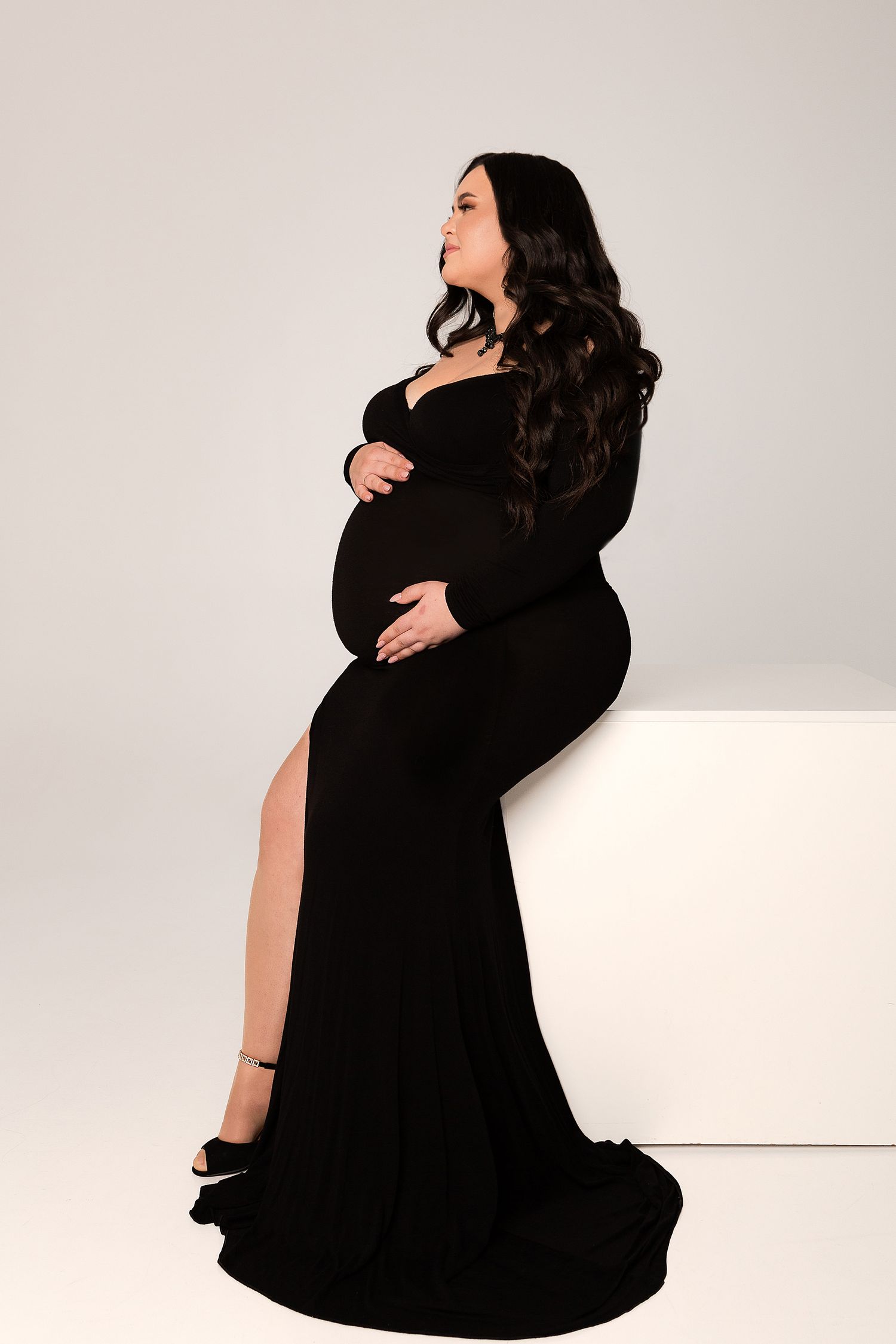 Elegant maternity portrait in a floor-length black gown with side slit on light background.