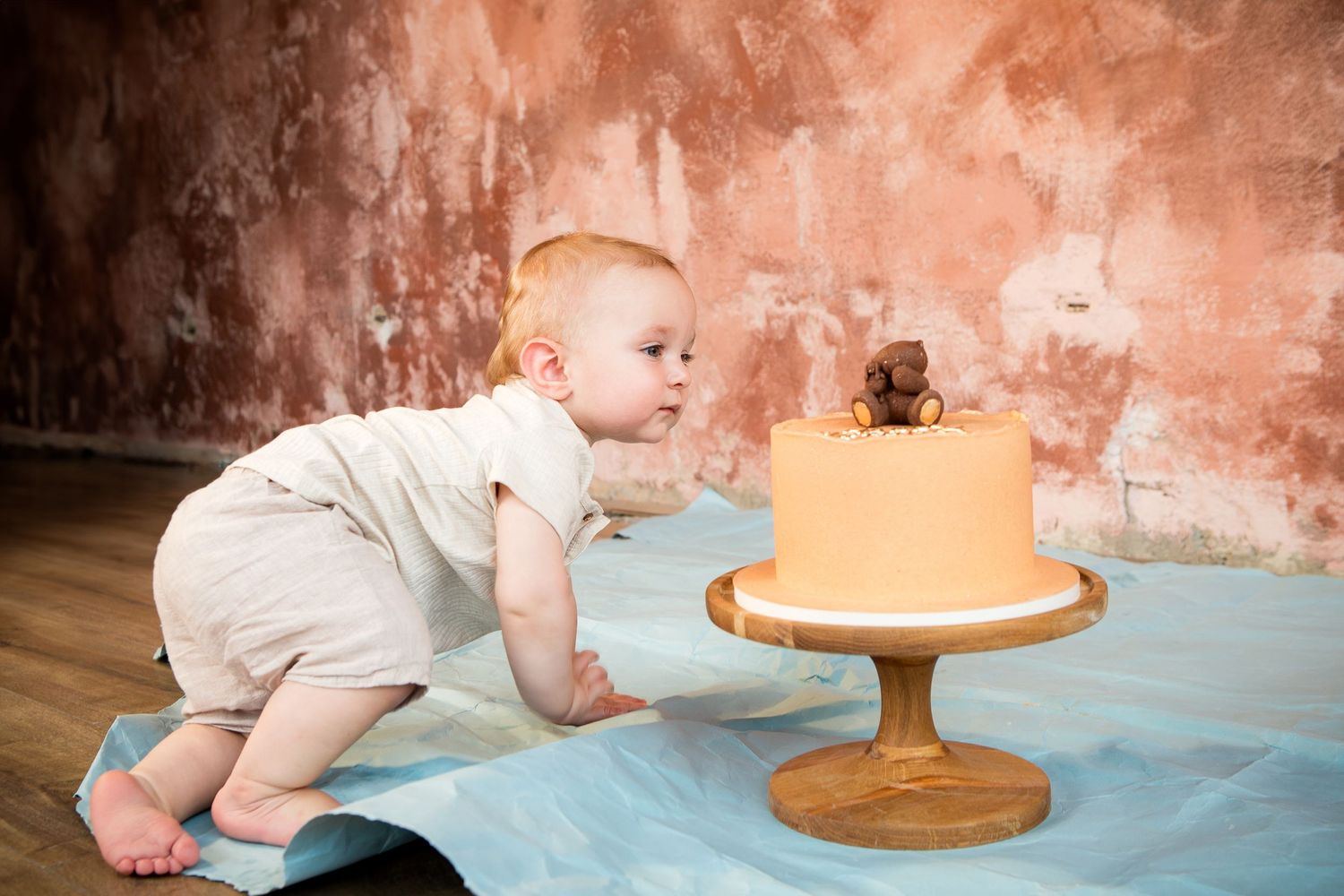 Baby in white outfit crawling towards orange birthday cake on blue blanket.