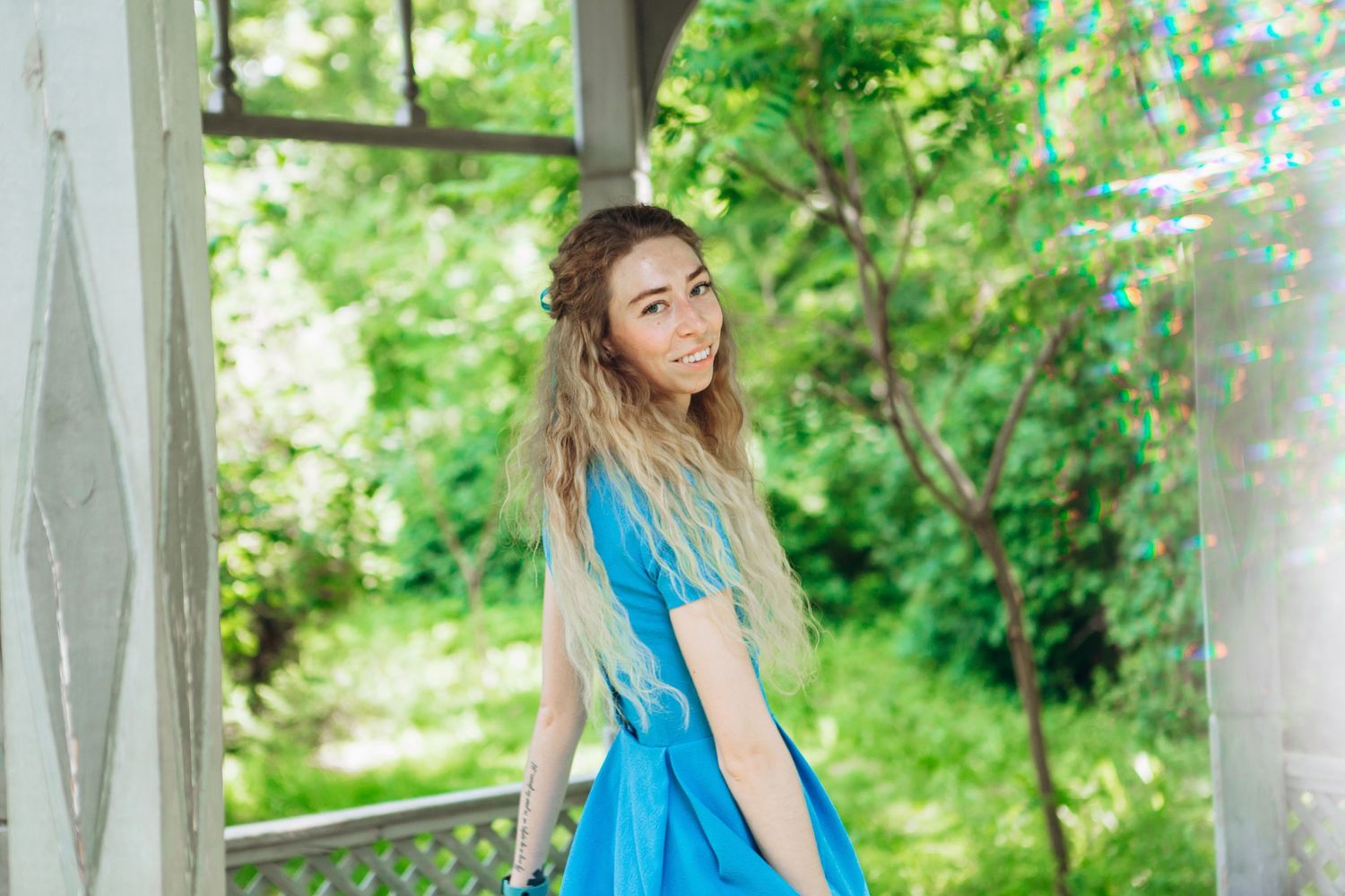 Woman in bright blue dress standing on a garden porch on a sunny summer day.