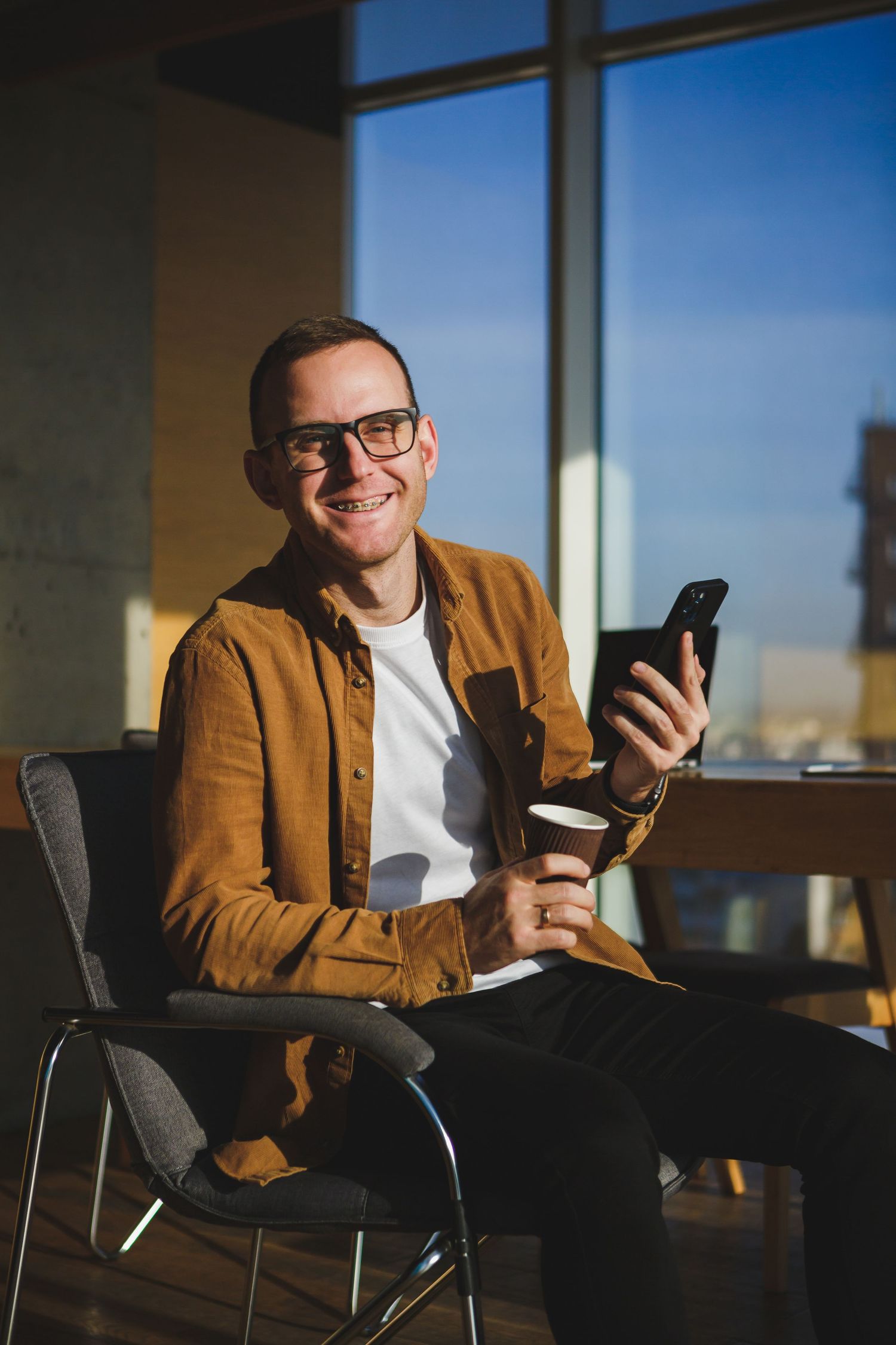 Professional in brown jacket smiling while using smartphone in sunny office setting.