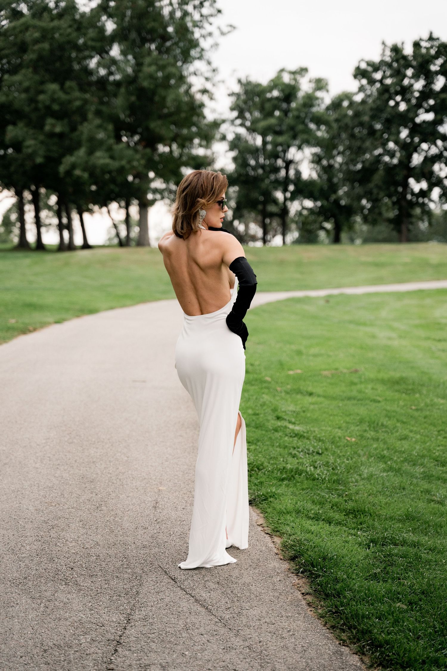 Backlit figure in backless white gown poses gracefully on golf course path.