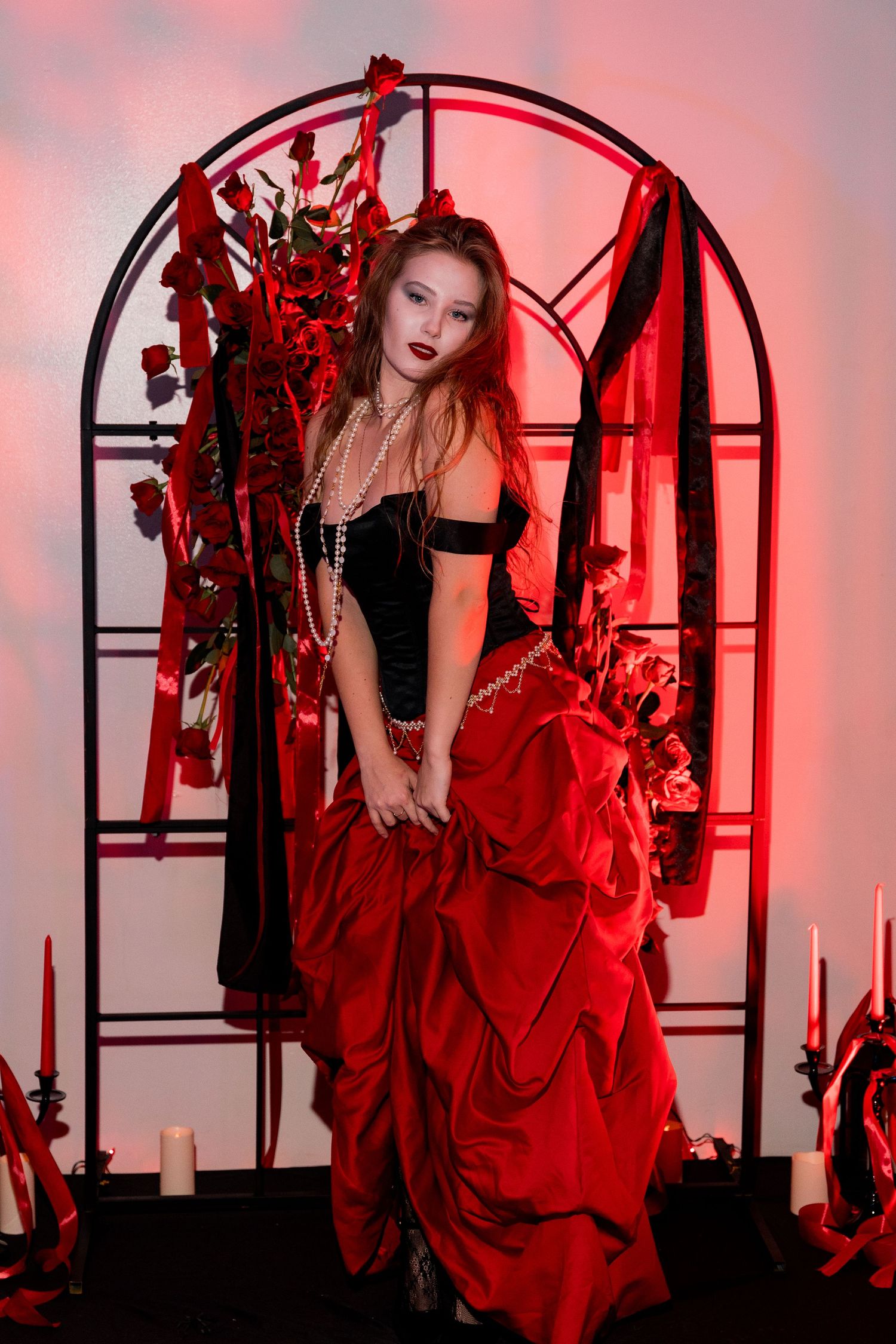 Woman in gothic dress poses dramatically in front of red-lit arch with Halloween decorations.
