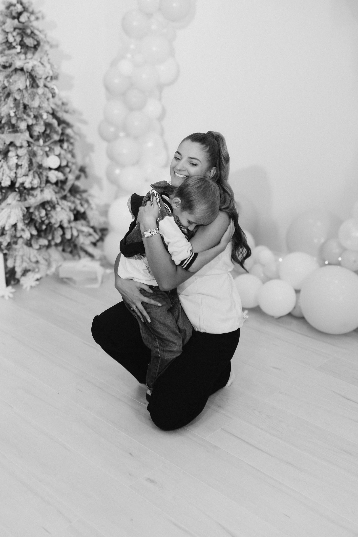 Black and white photo of a tender embrace in front of a Christmas tree with balloon decorations.