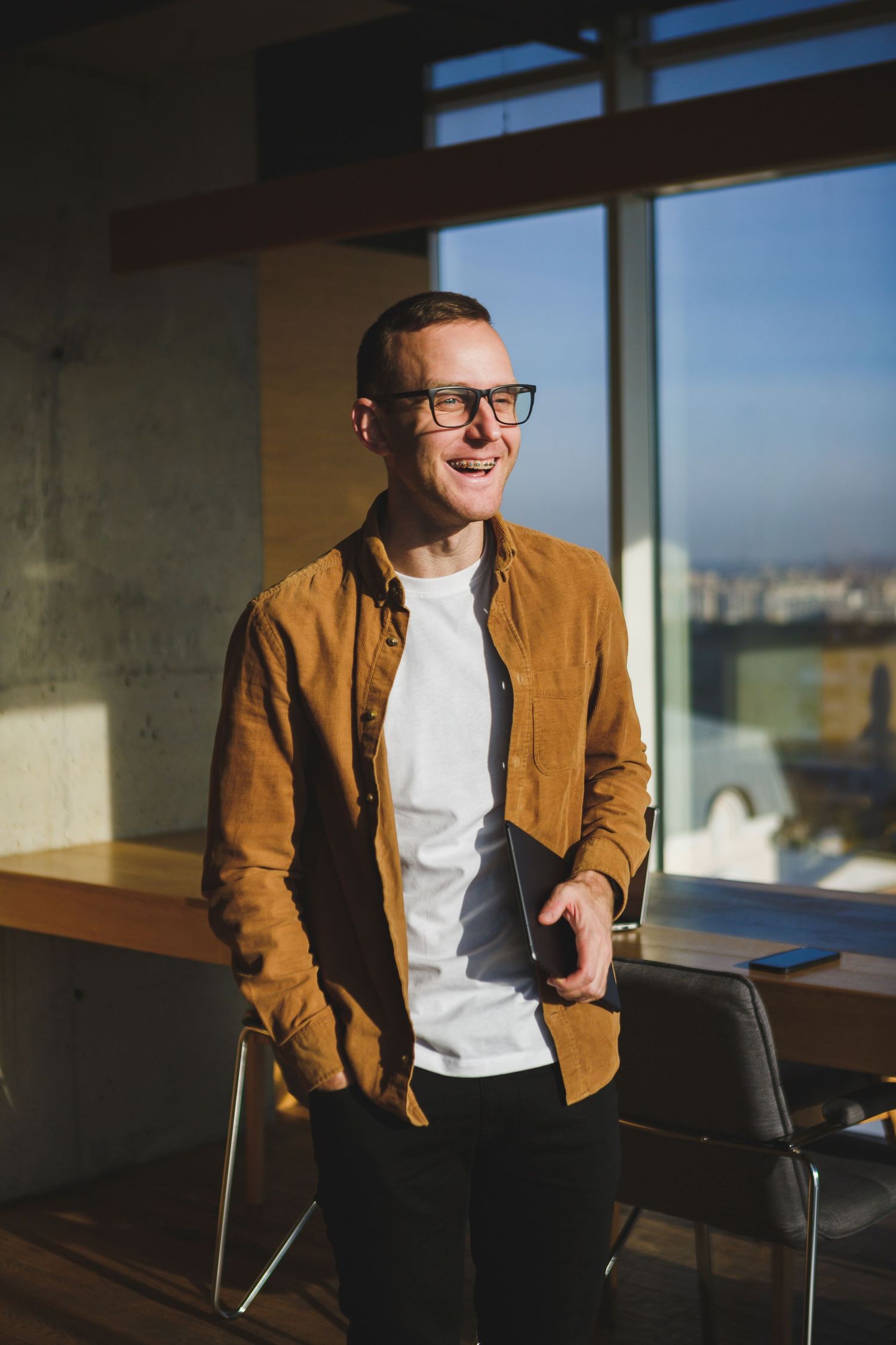 Professional in brown jacket and glasses stands confidently by window in modern office space.