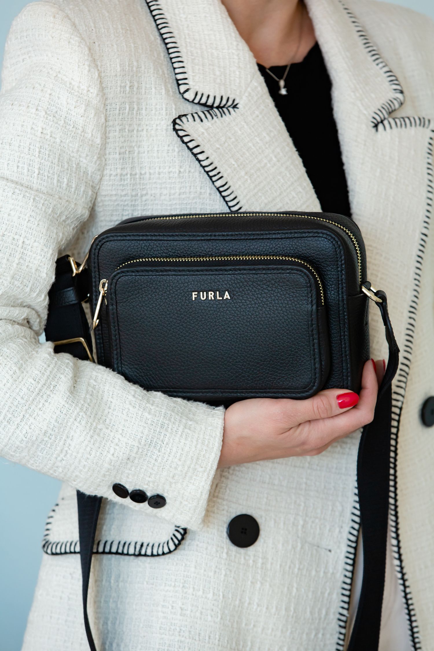 Black Furla camera bag held against white contrast-stitched blazer.