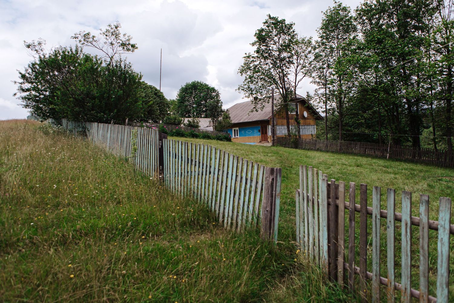 Rural countryside house behind weathered wooden fence on grassy hillside.