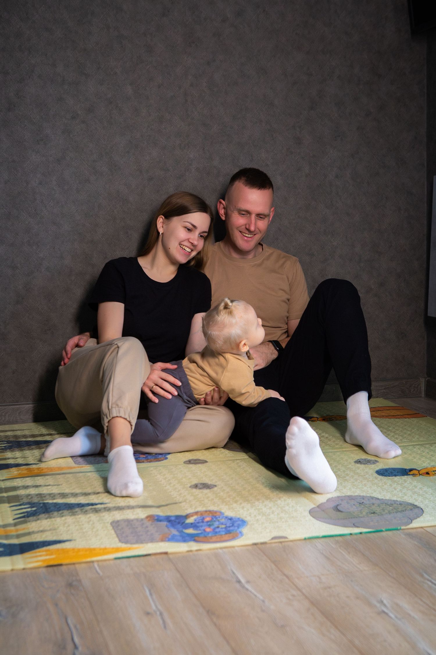 A couple relaxes together on the floor against a gray wall while wearing casual clothing.