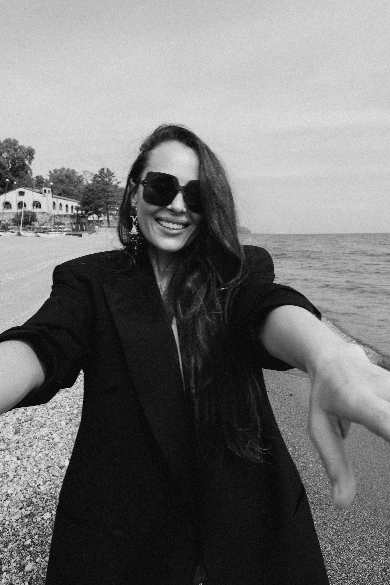 Black and white photo of someone in sunglasses and black coat smiling at a beach with buildings in background.