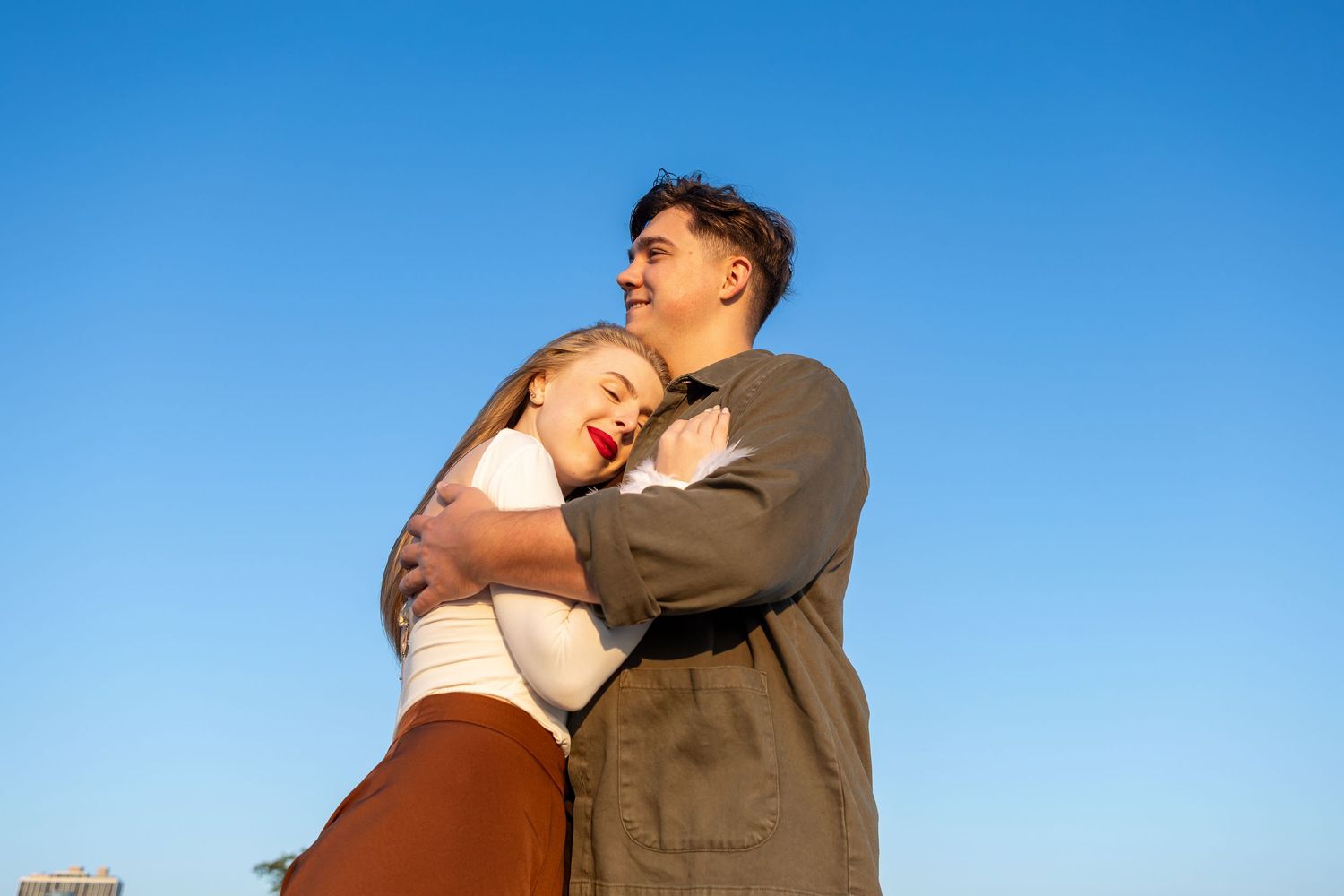 A couple embraces outdoors against a bright blue sky during golden hour lighting.