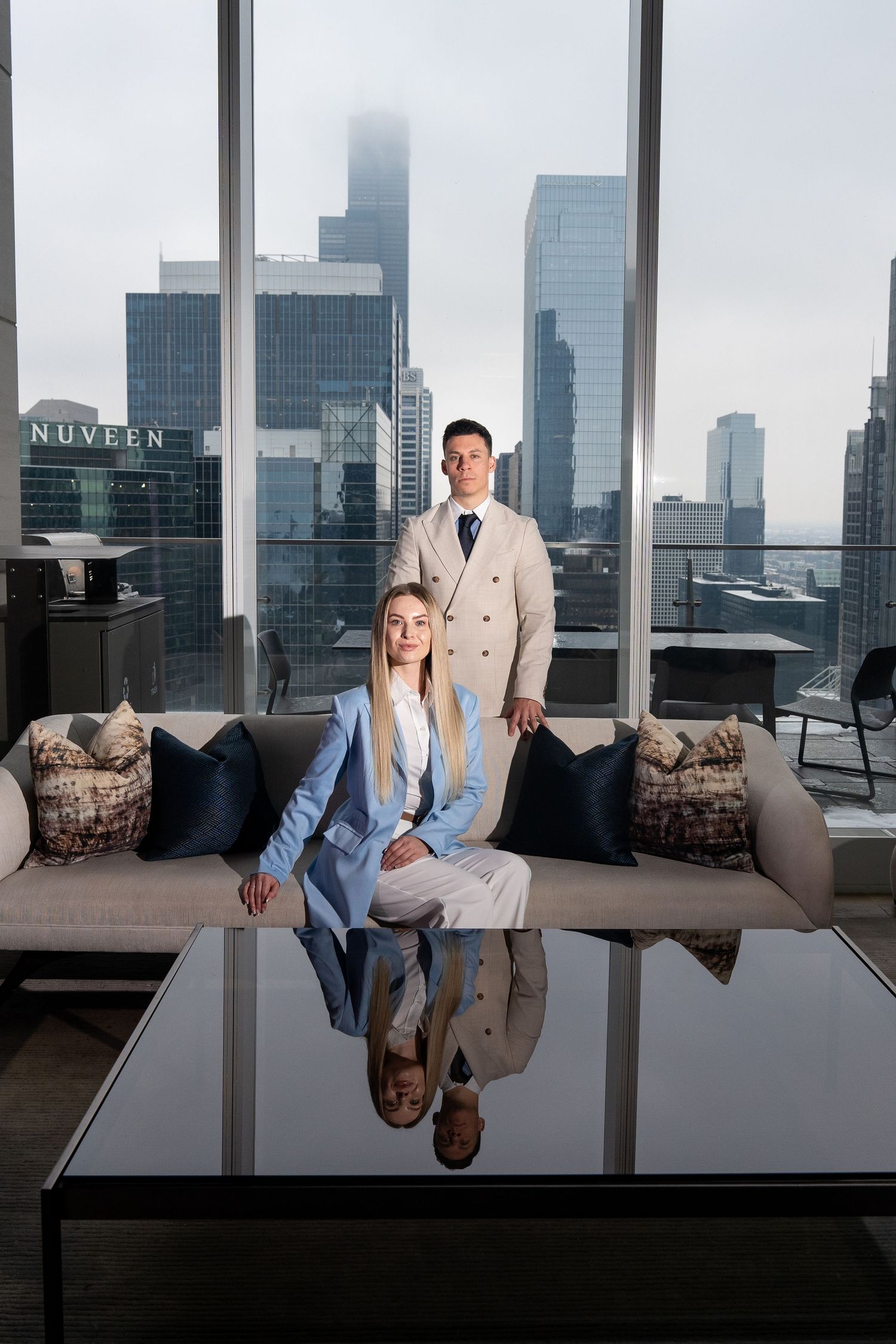 Two professionals in contrasting suits pose in modern office setting with dramatic city views.