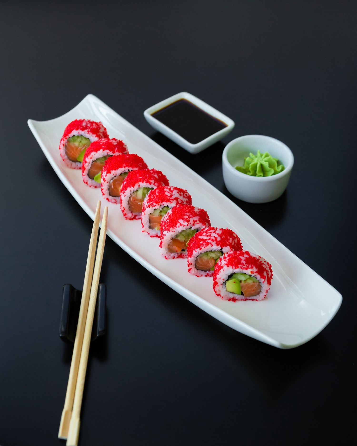 Fresh sushi roll with bright red tobiko displayed on white plate with soy sauce and wasabi accompaniments.