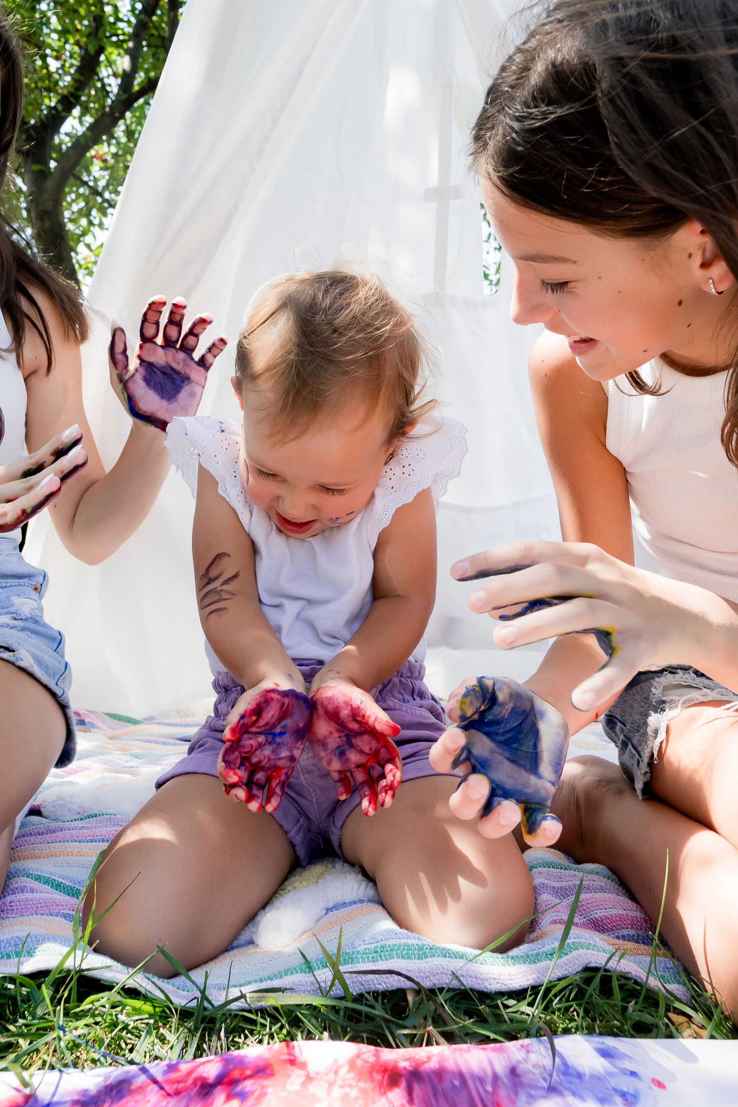 Group enjoying finger painting outdoors on a white blanket under trees.