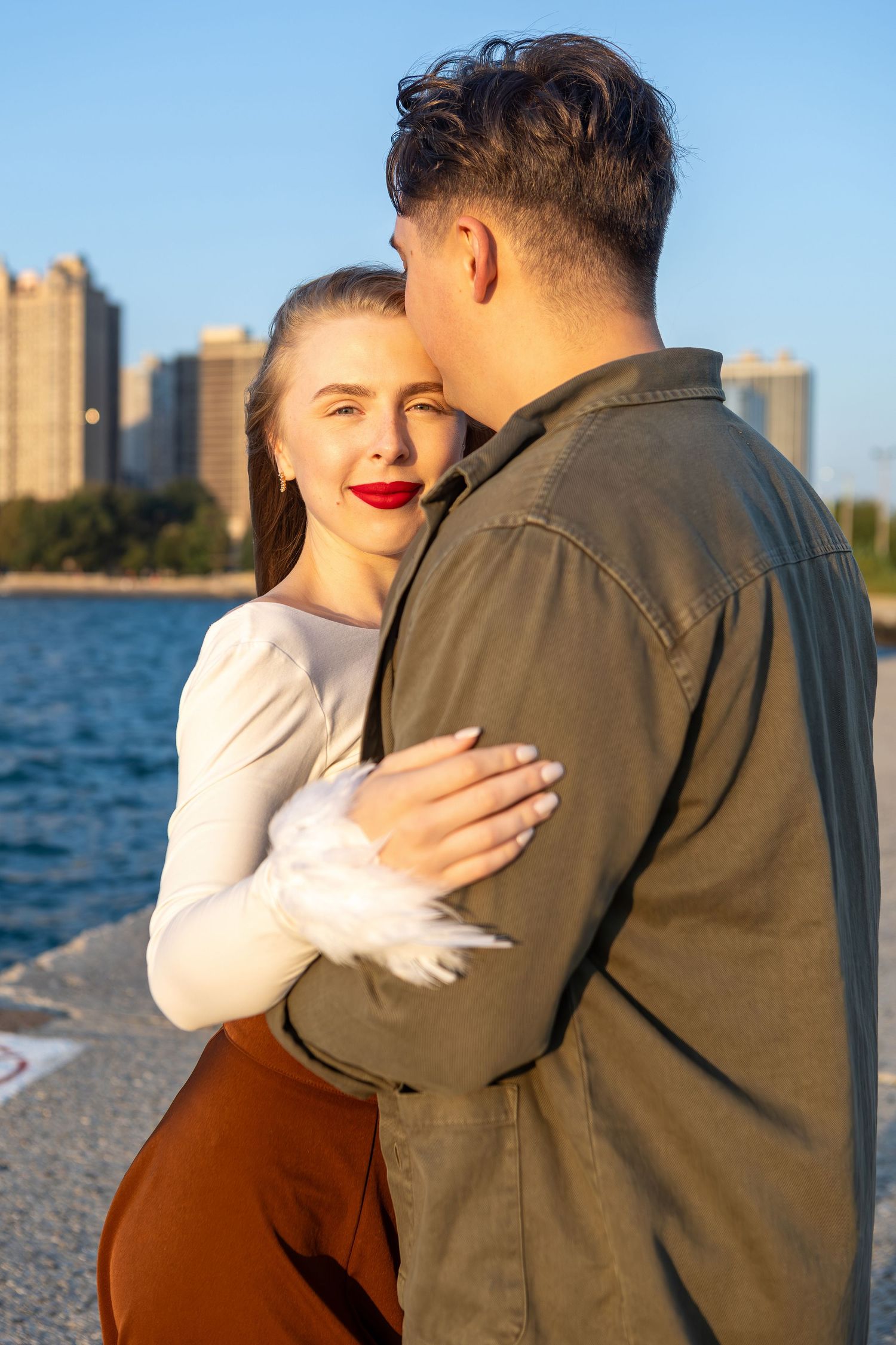 A couple embraces by the waterfront at sunset with city buildings visible in the background.