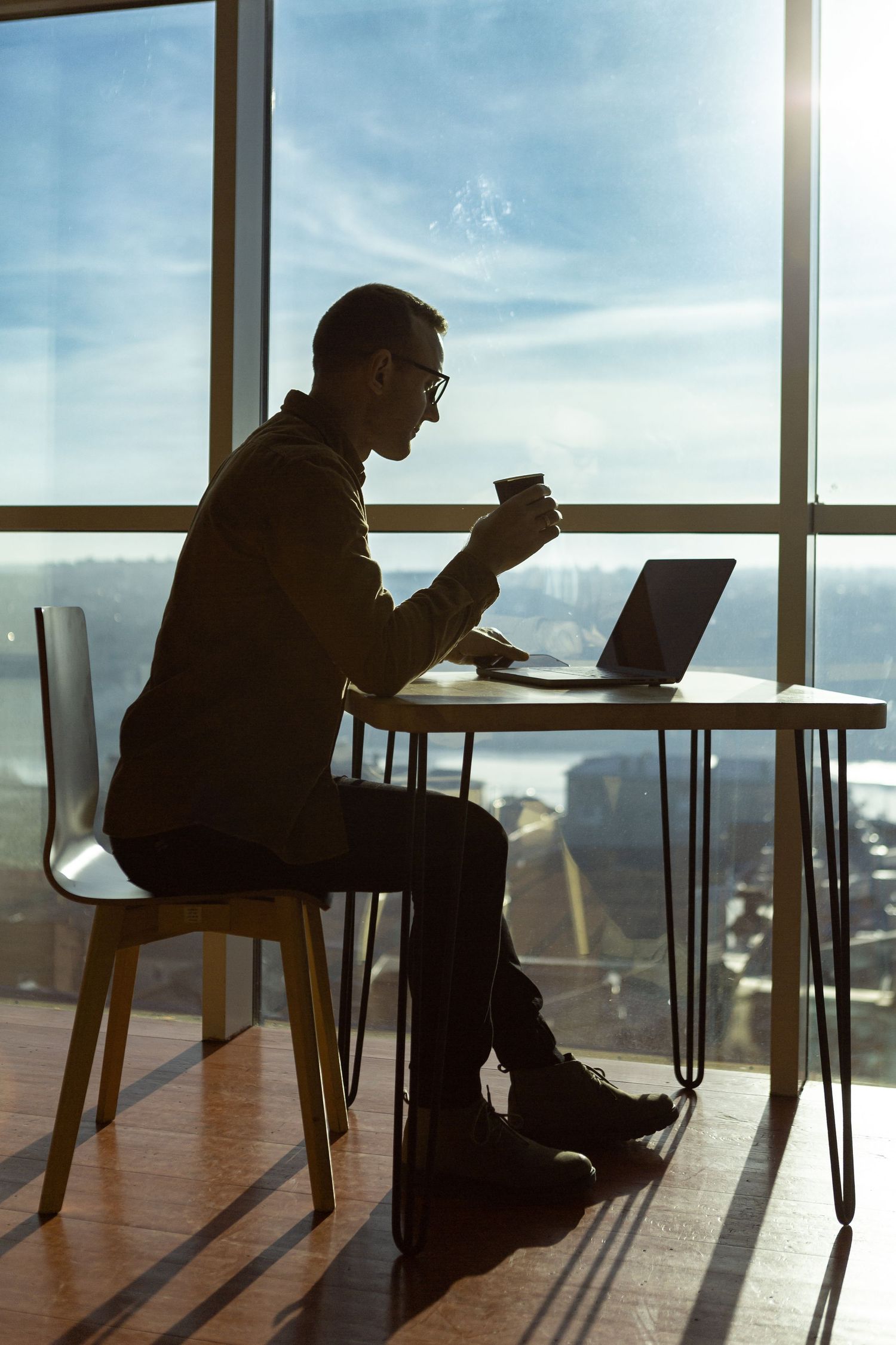 Silhouette of professional working on laptop by window with coffee cup in modern office space.