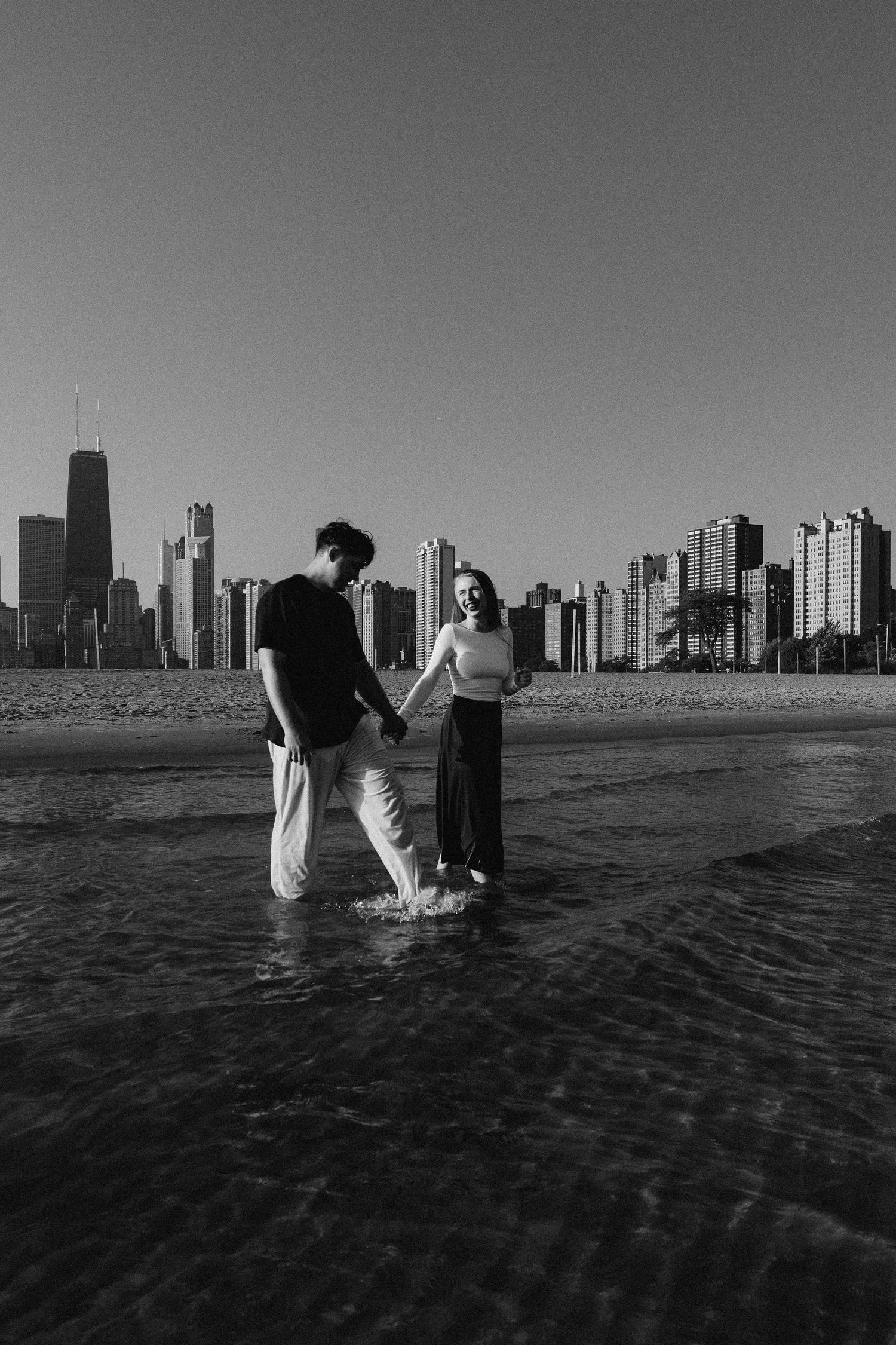 A couple holds hands at North Avenue Beach with Chicago's iconic skyline silhouetted against a clear sky in black and white.