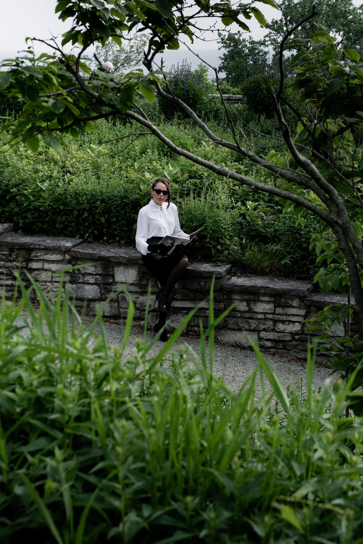 Person in white shirt sits on stone wall surrounded by lush greenery and trees in outdoor garden setting.