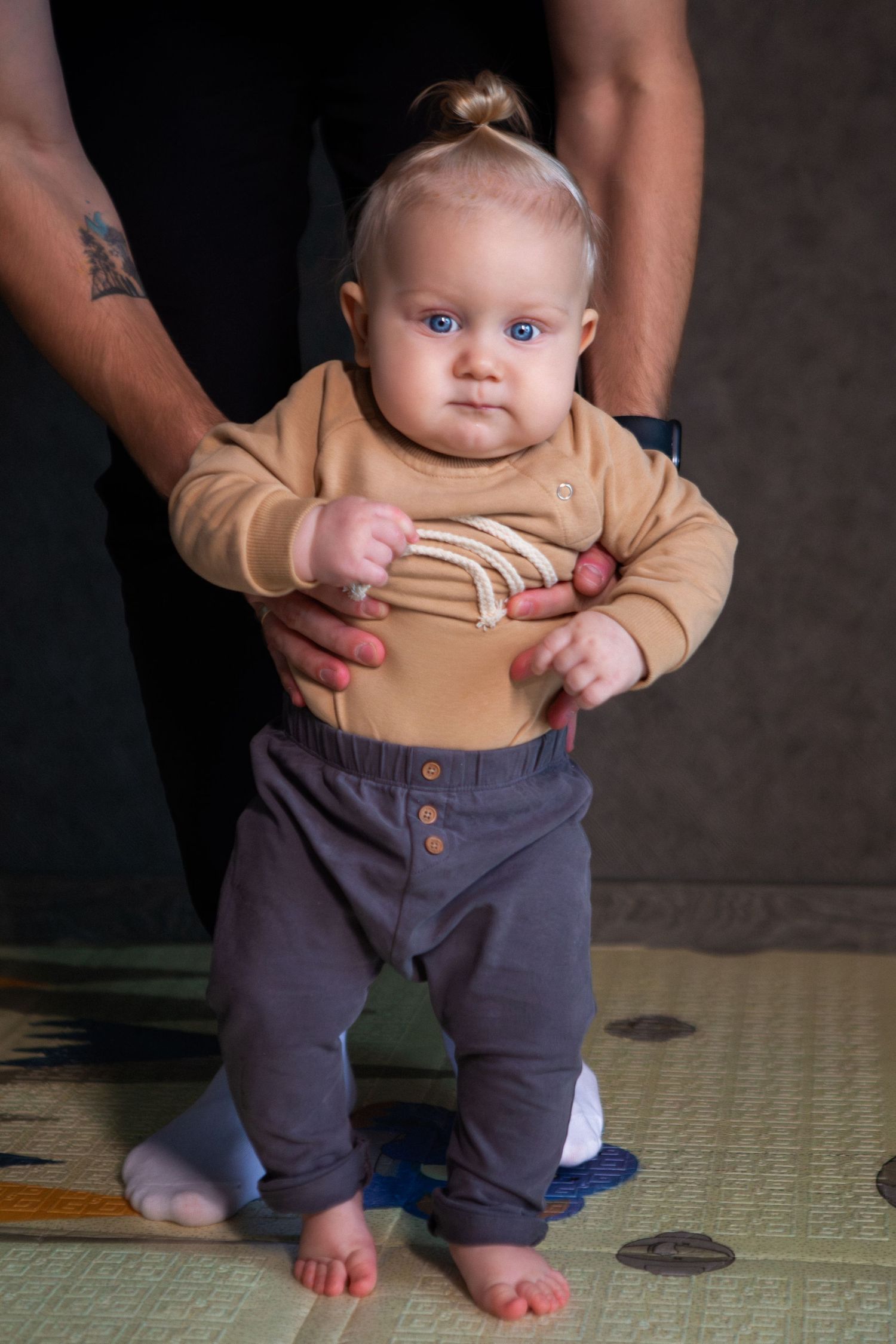 A series of photos showing a baby in tan sweater and navy pants being held while standing on a green mat.