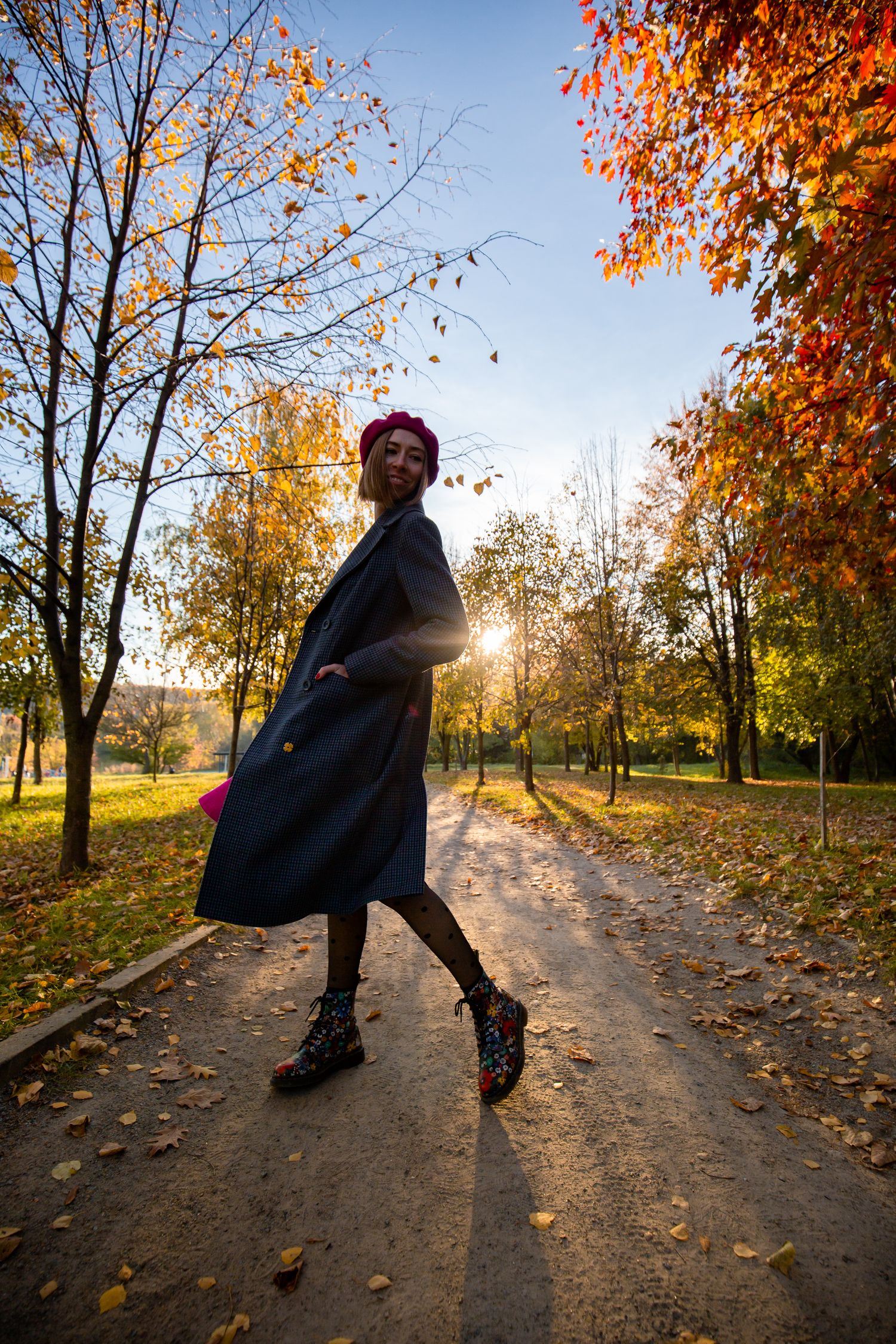 Figure in dark coat twirling on park path surrounded by fall foliage.