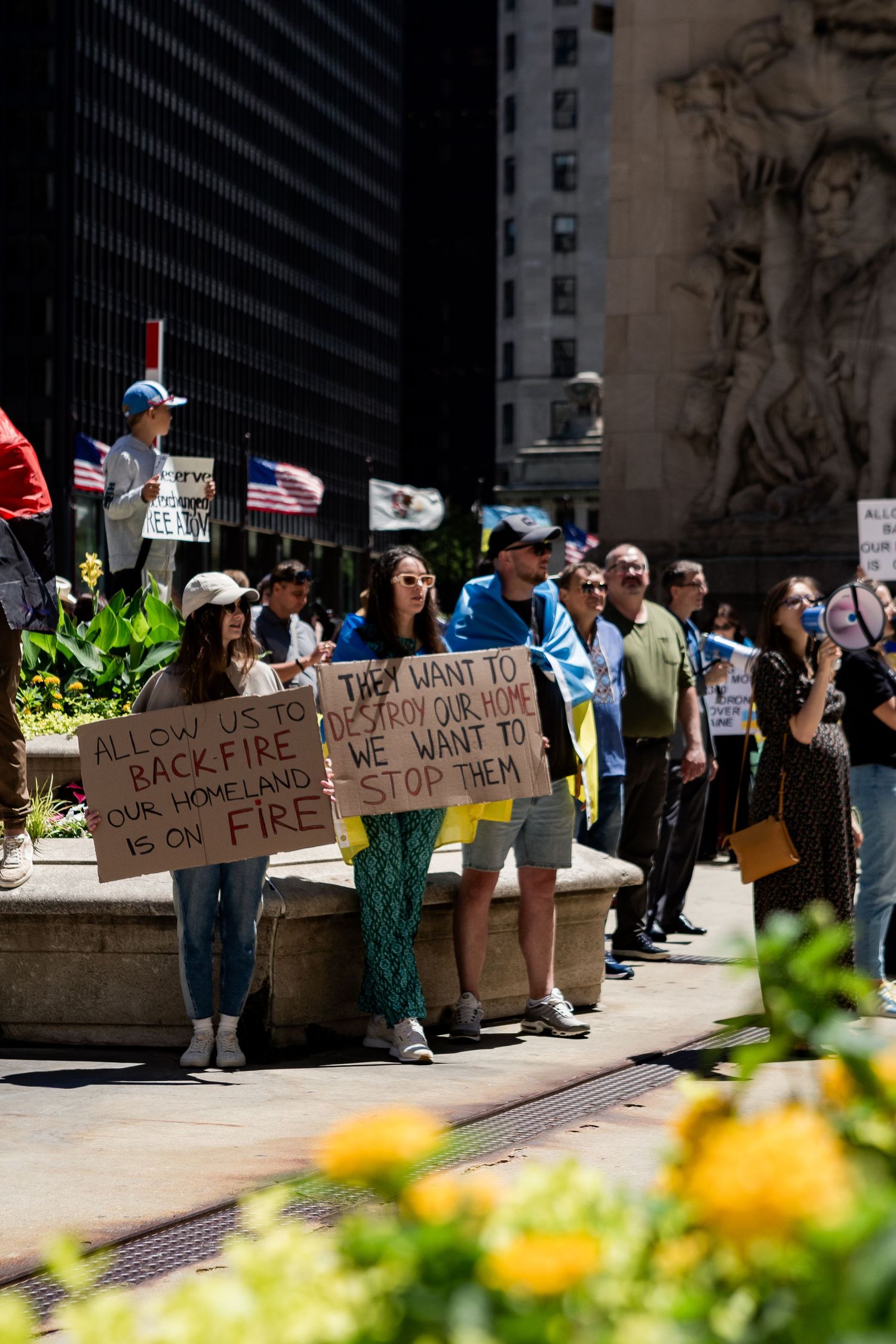Protesters line up with signs along urban street with flowers and buildings in background.