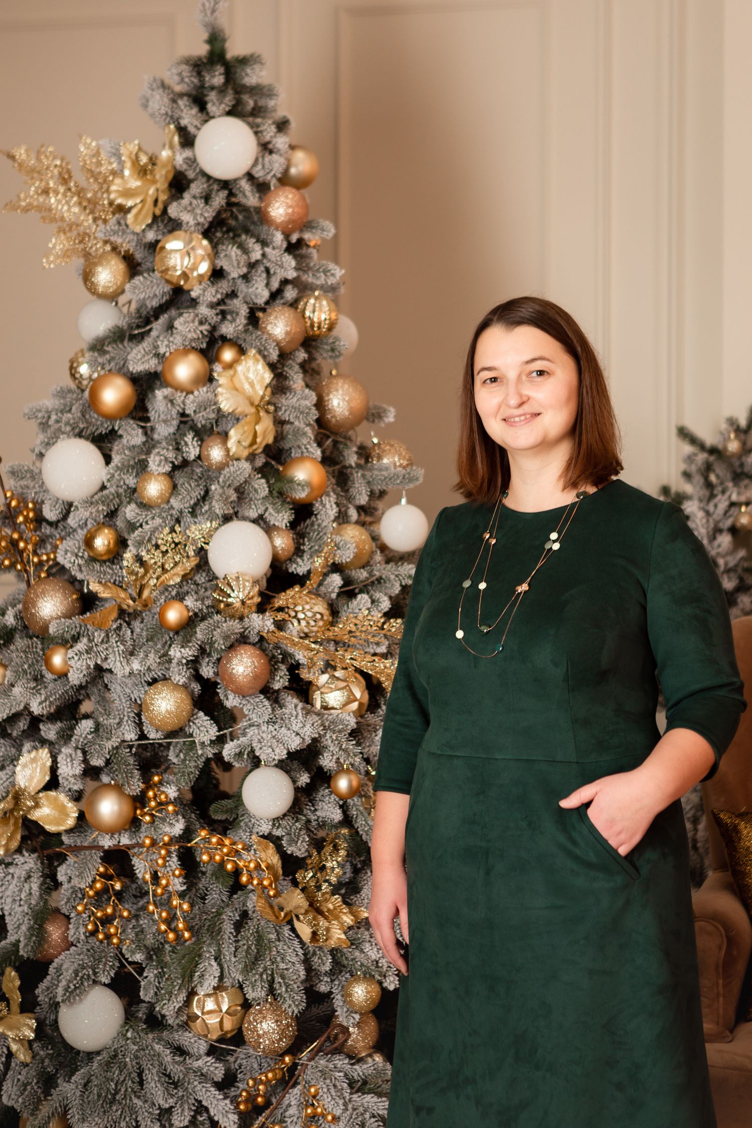Woman in emerald green dress stands beside elegantly decorated Christmas tree with gold and white ornaments.