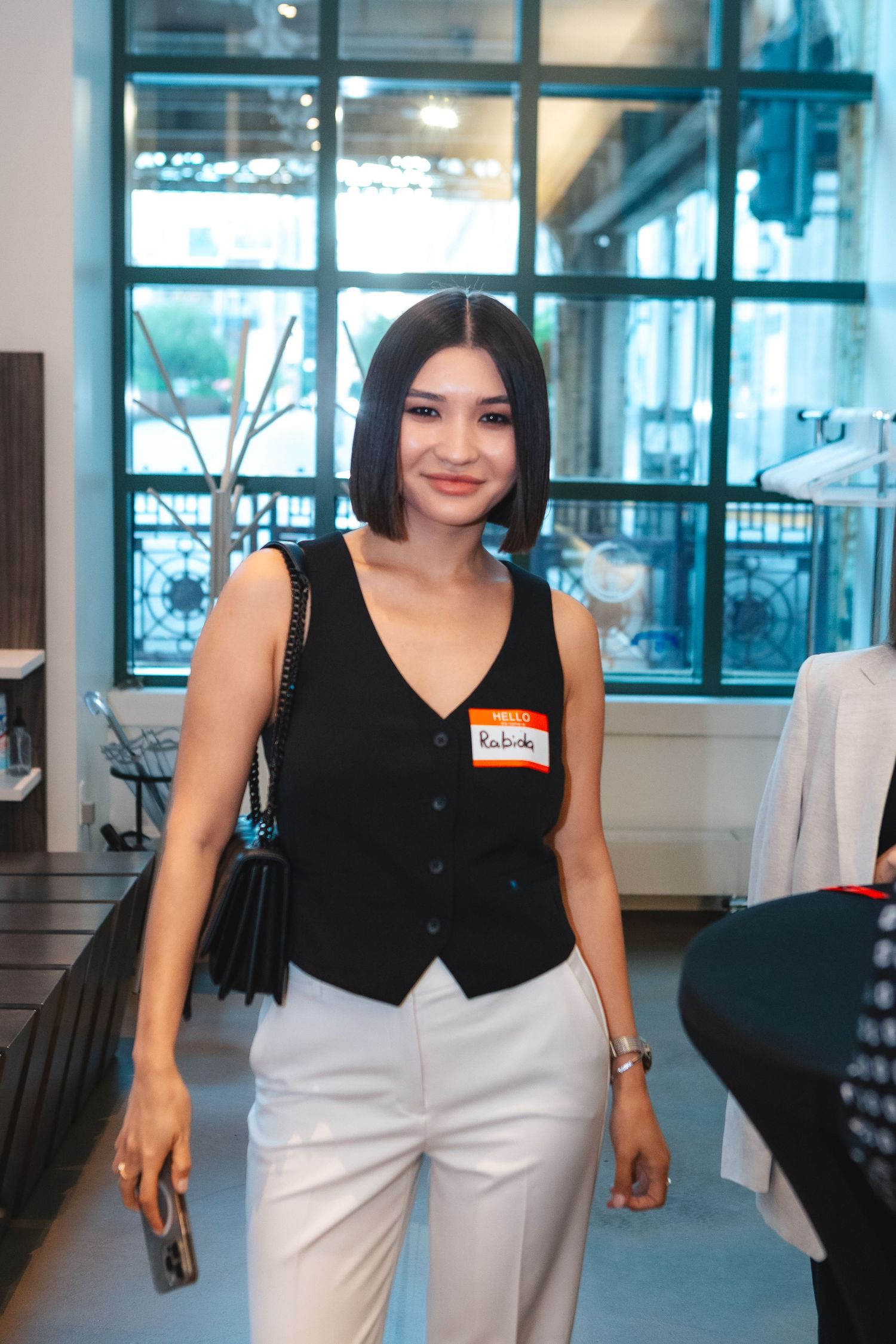 A woman wearing a black vest and white pants stands in a modern office setting with glass windows.