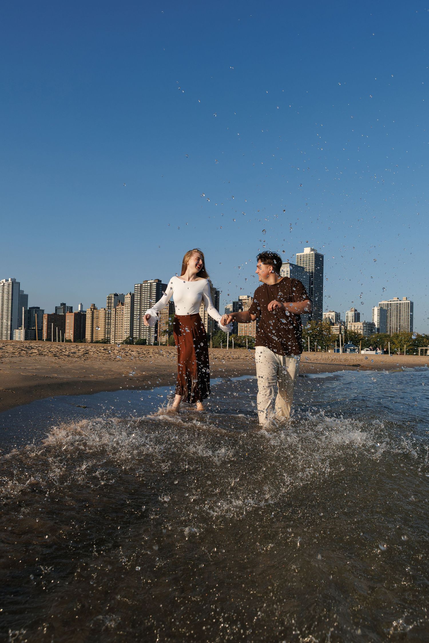 People playing in ocean waves at sunset with city skyline visible in background.