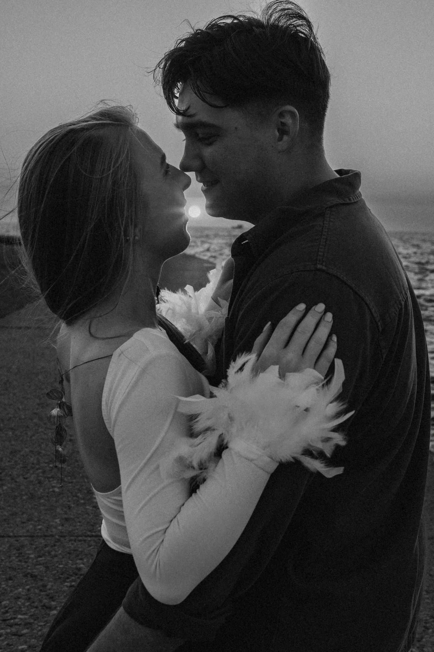A romantic black and white portrait of a couple embracing outdoors with soft lighting and ocean in background.